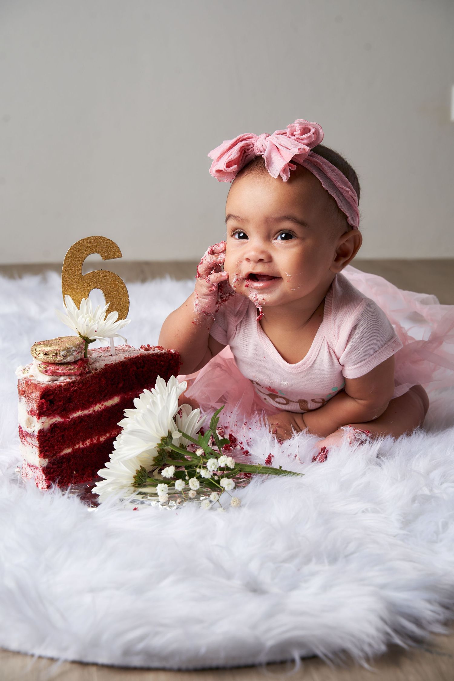 Baby in pink outfit lying on white fur with red velvet cake and white daisies during 6 month milestone photo session.