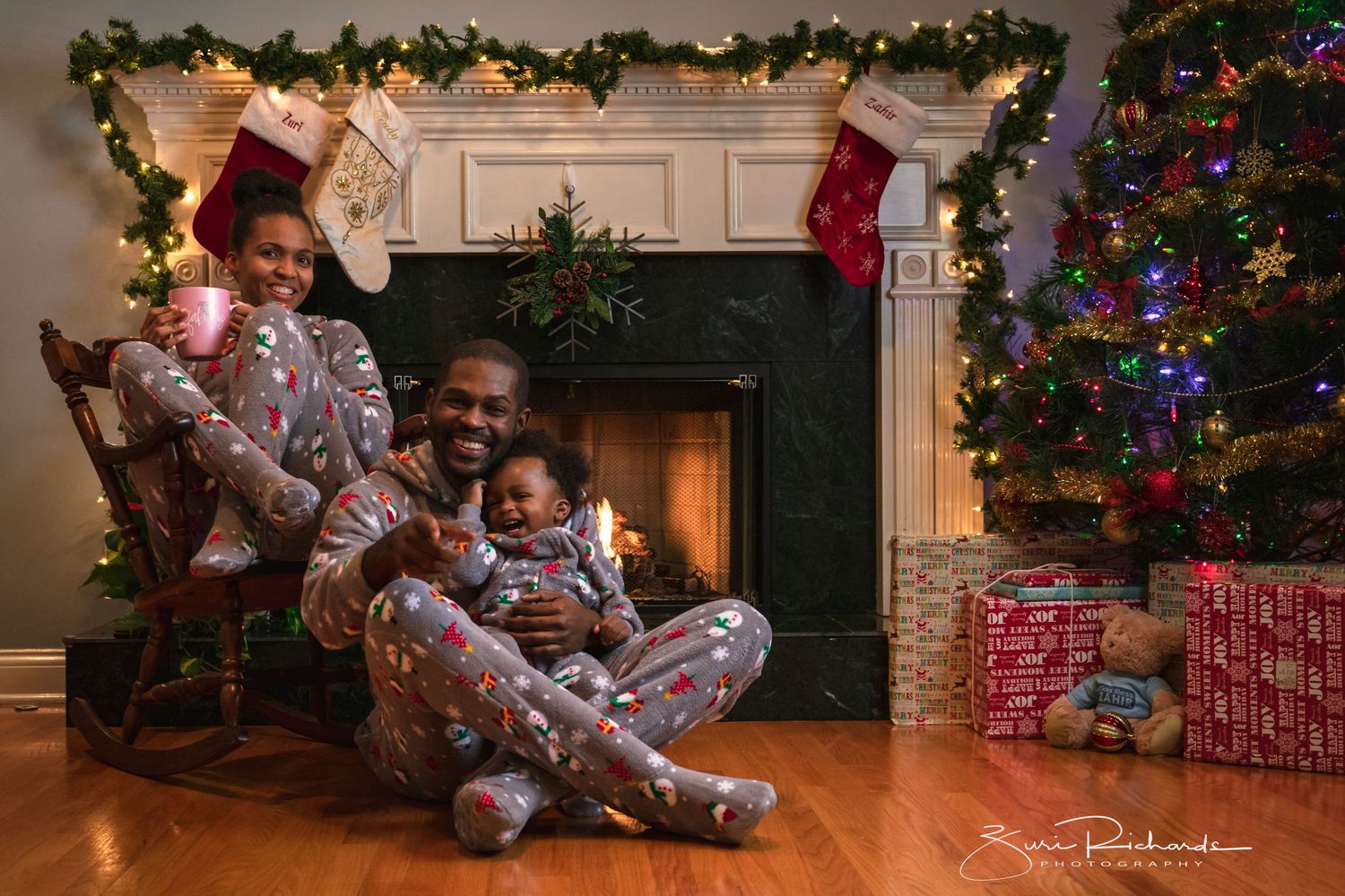 A cozy Christmas scene with a child sitting in pajamas near a decorated tree and stockings hung by the fireplace.