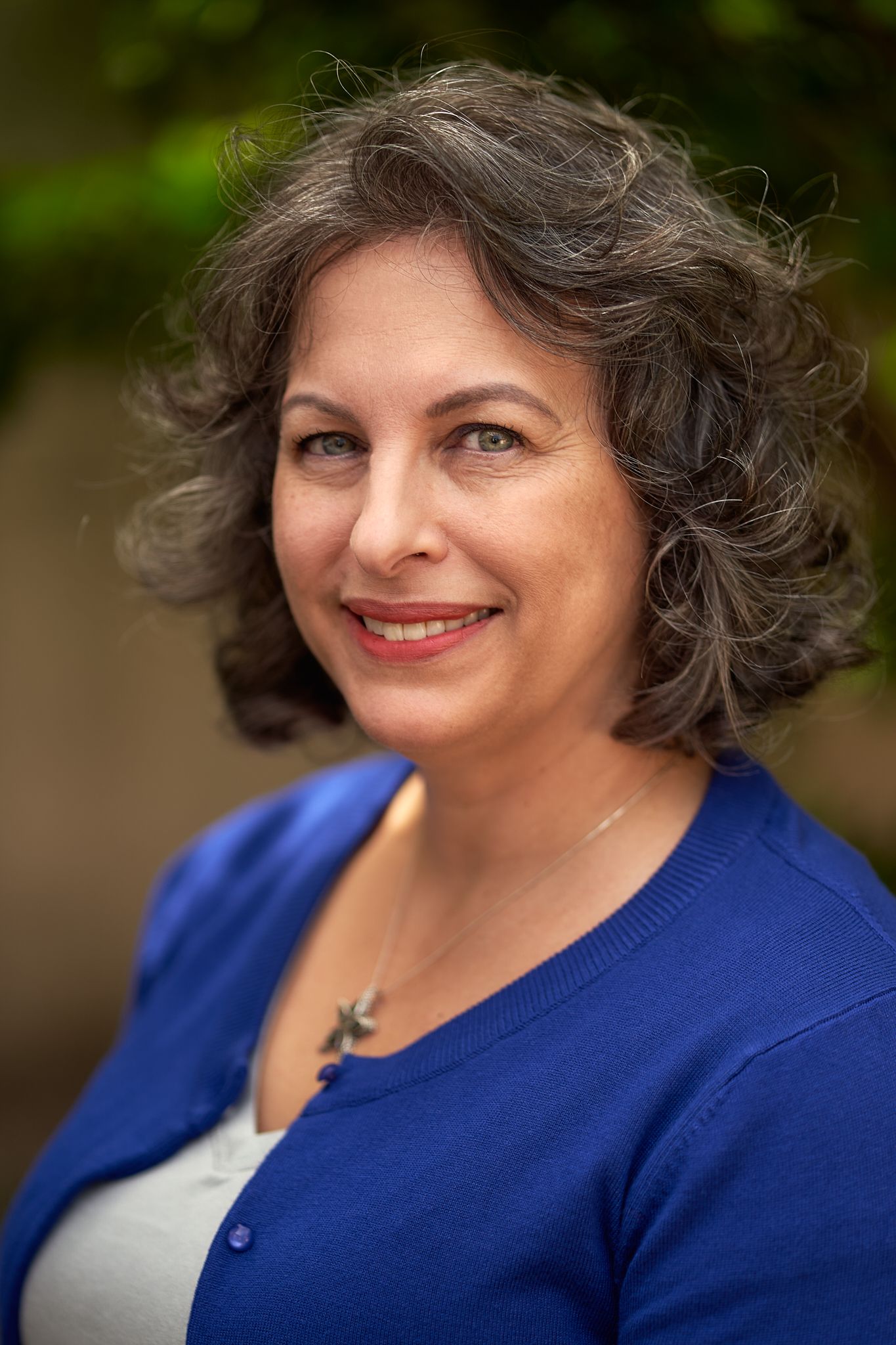 Woman in blue cardigan and white top smiling warmly in outdoor portrait series.
