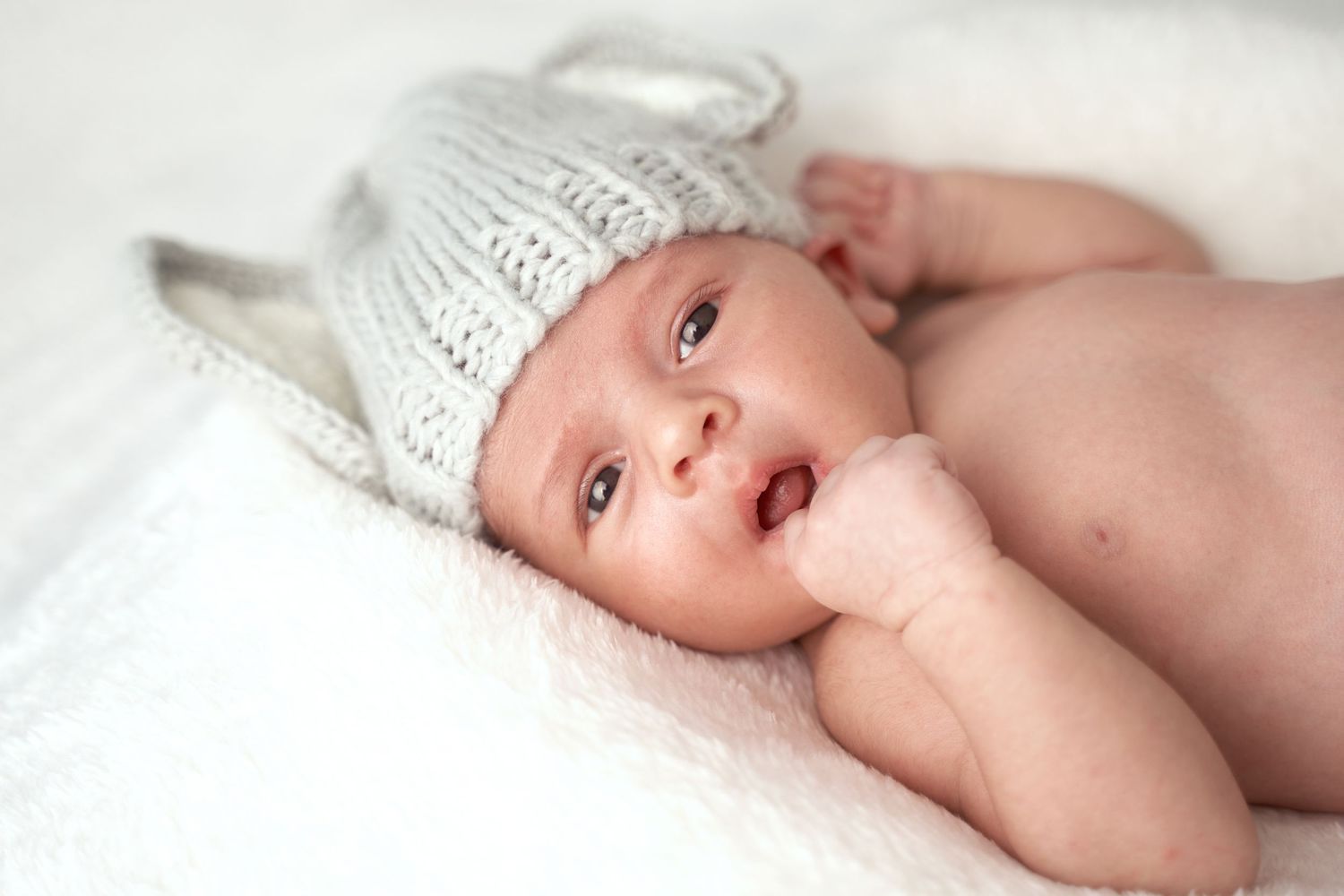 A newborn baby lies on soft white bedding while being gently touched by caring hands.