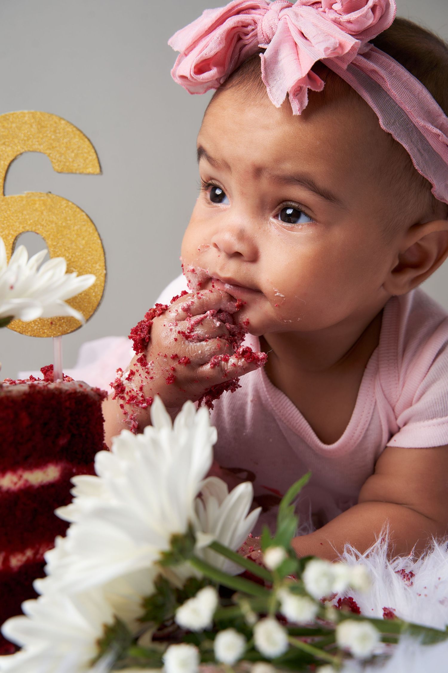 A baby in pink with a pink bow enjoys red velvet cake next to white daisies and a gold number 6 decoration.