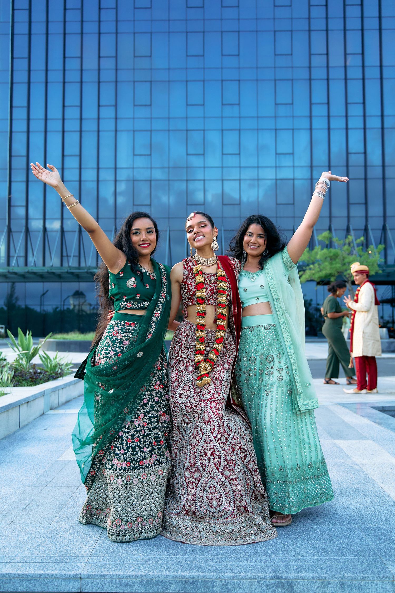 Three women in colorful Indian lehengas pose joyfully against a glass building backdrop.
