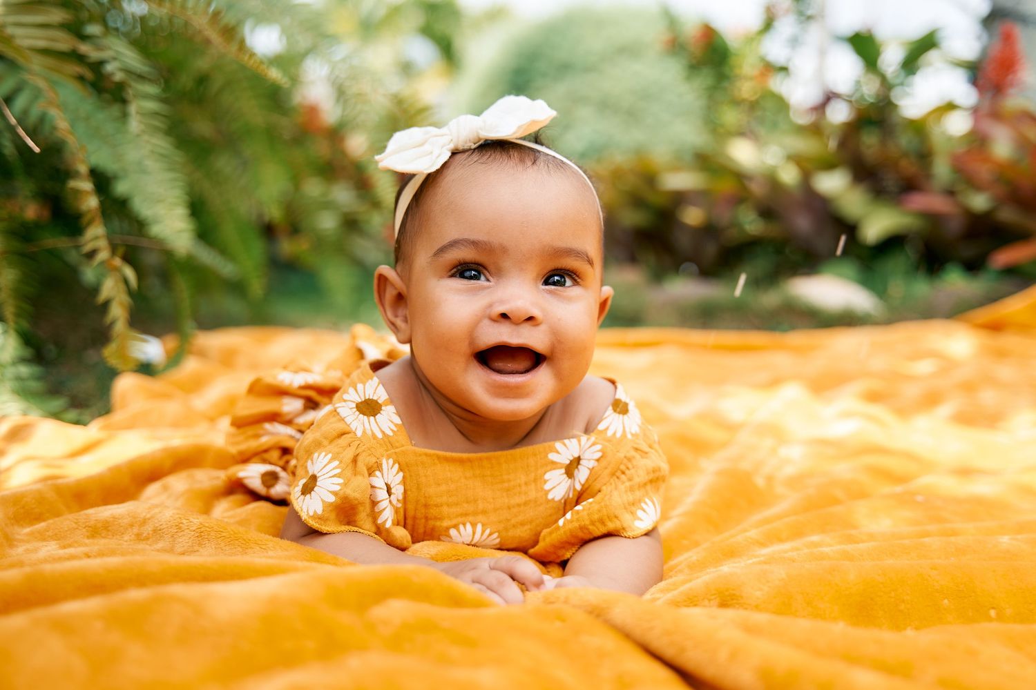 Baby in white outfit smiles while lying on orange blanket outdoors with autumn foliage visible.