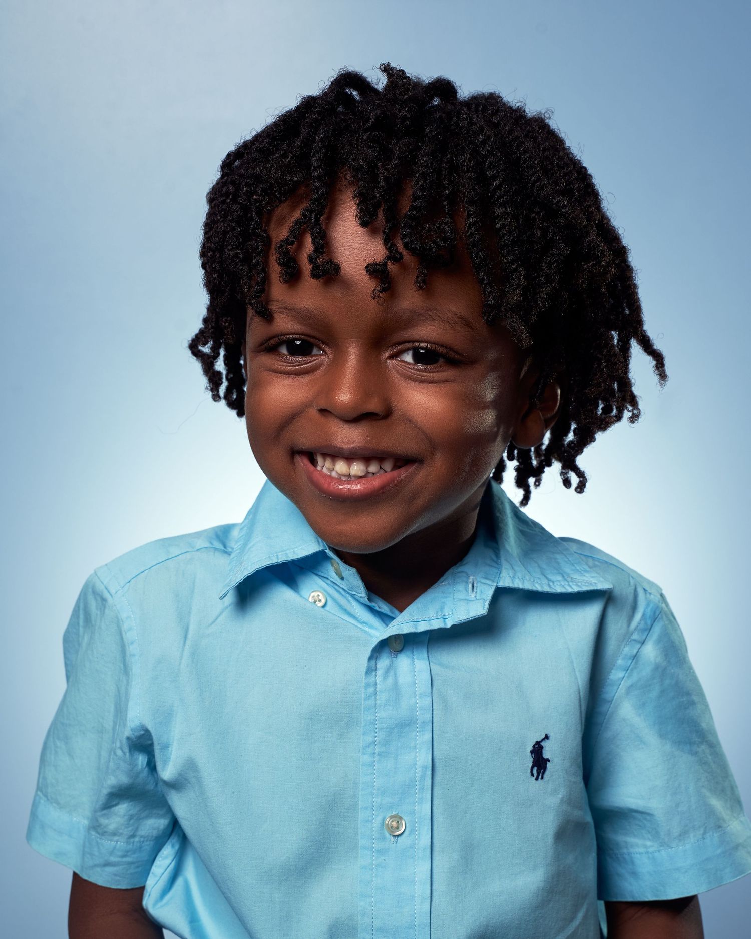 Young child wearing a light blue button-down shirt smiles while posing against a blue background.