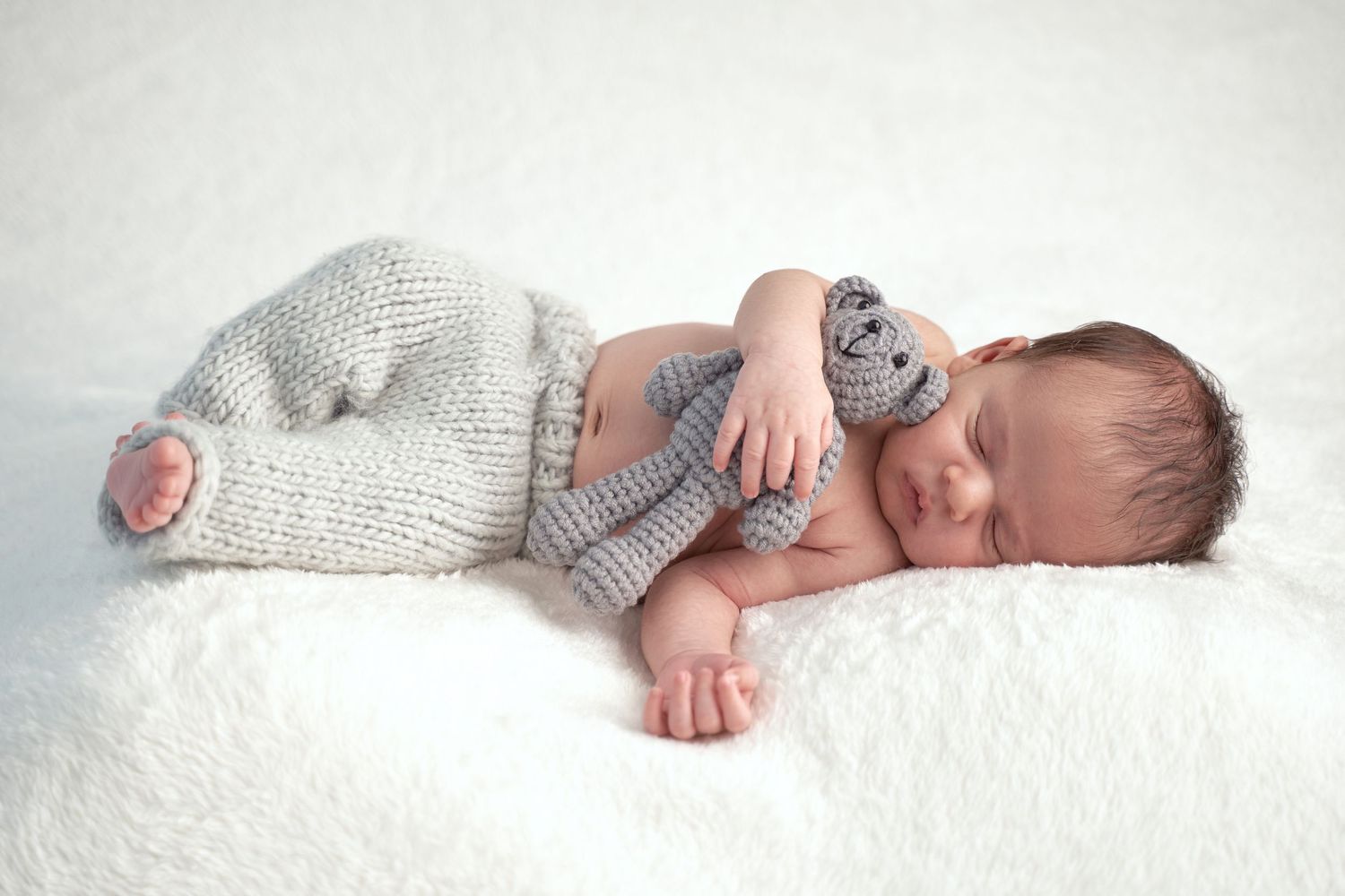 A sleeping baby in a gray knit sweater lies on a soft white blanket in a peaceful pose.