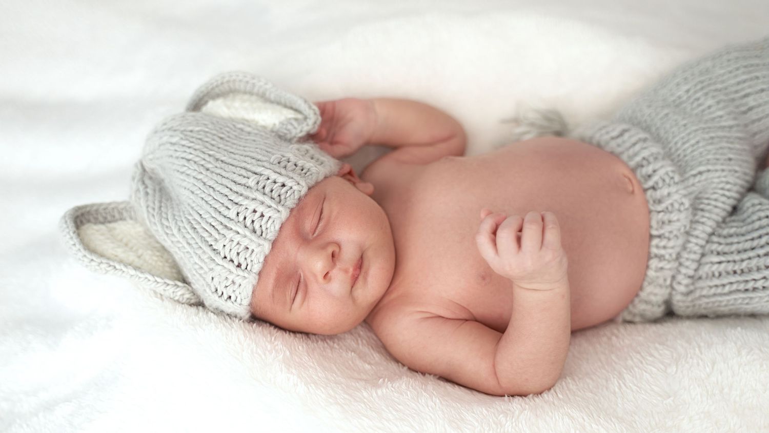 A newborn baby wearing a white knit hat lies on soft white blankets in a peaceful sleeping pose.