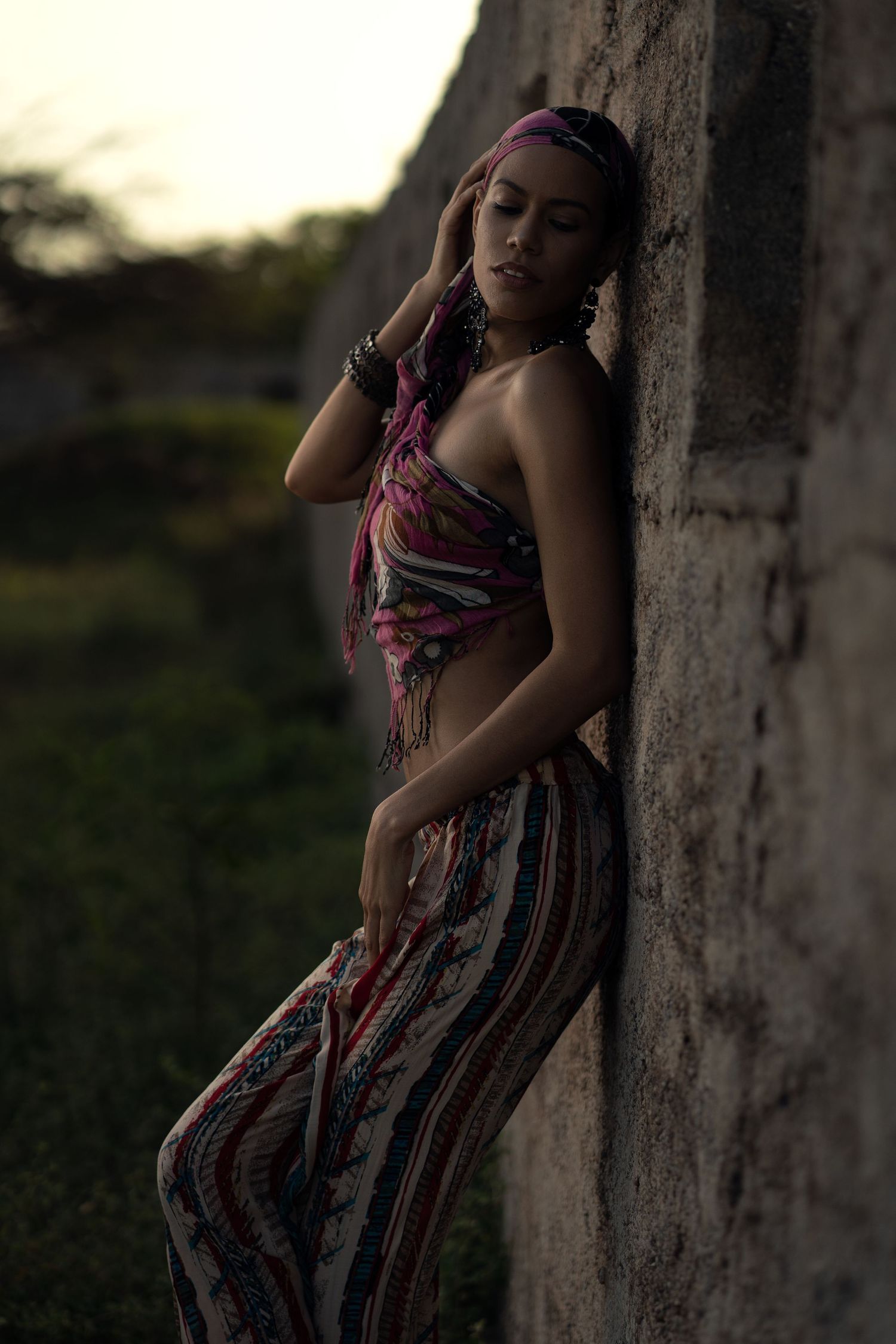 Artistic portrait of a person in patterned clothing leaning against a wall at sunset.
