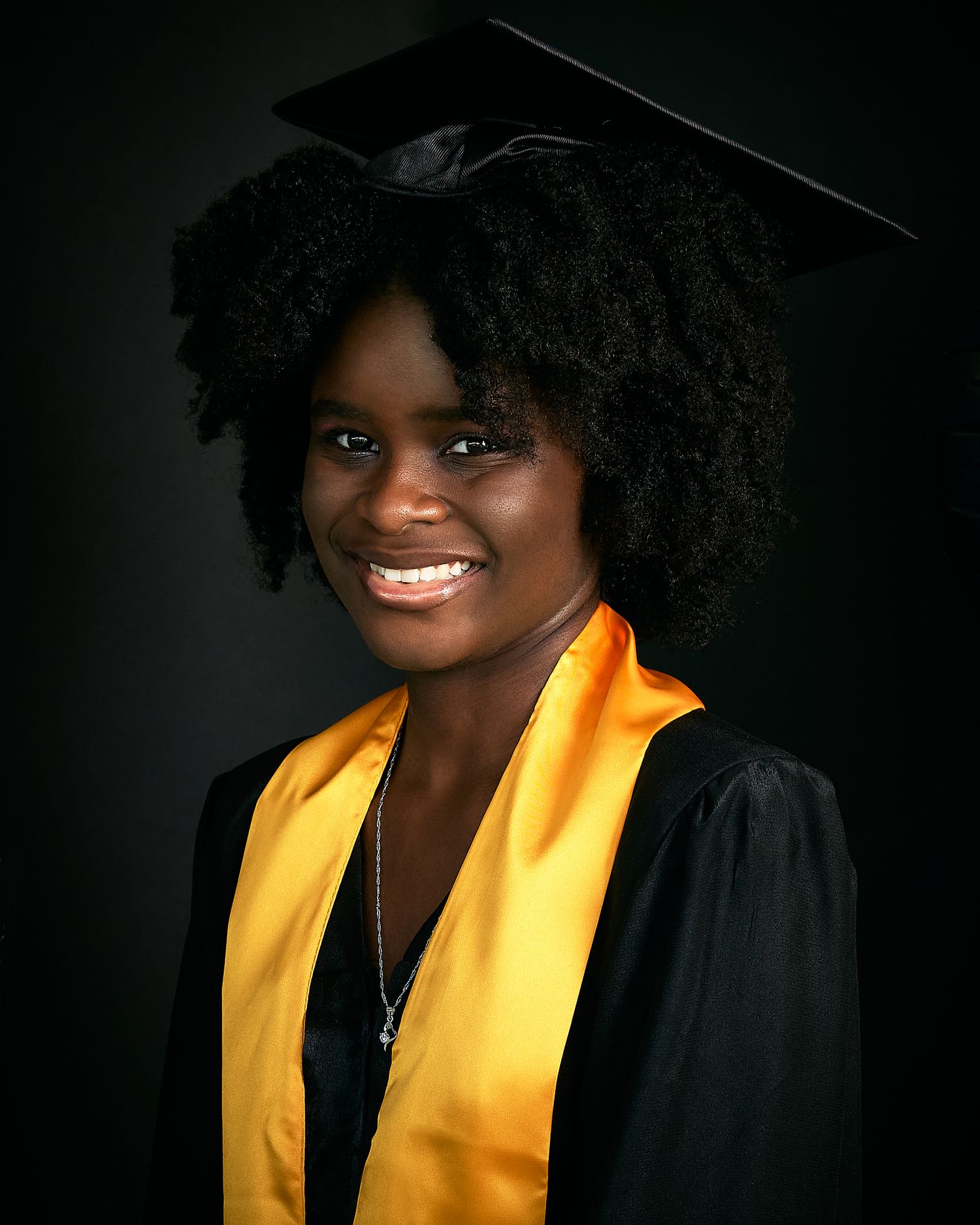 Graduate in black cap and gown with gold stole smiling brightly against dark background.