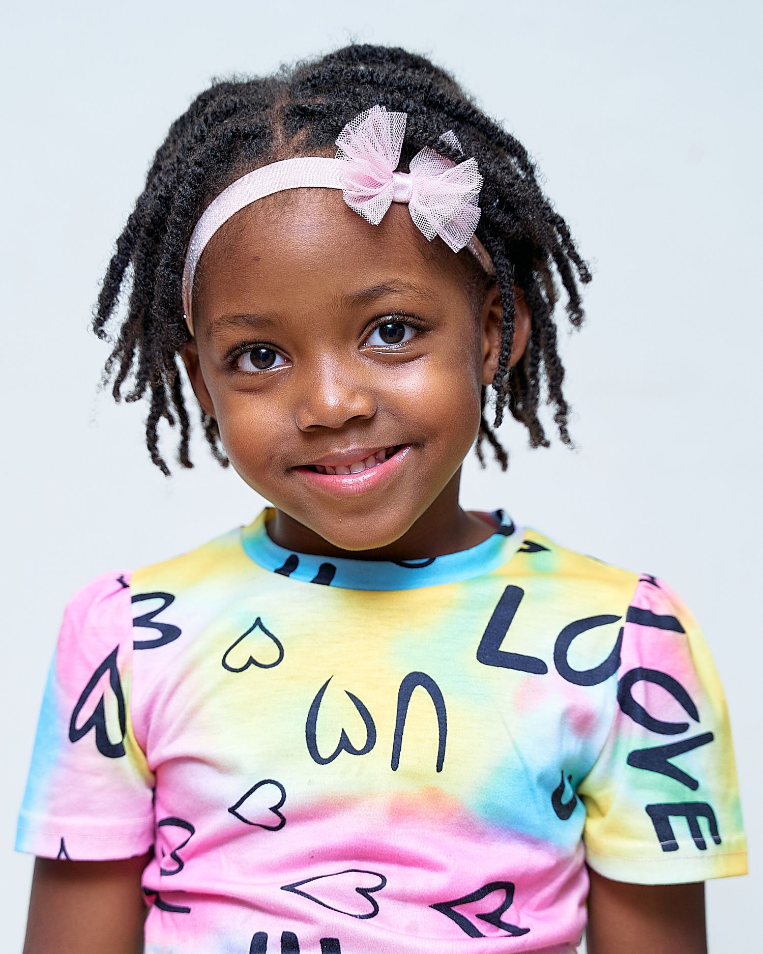 Child wearing colorful tie-dye shirt with heart patterns poses against white background.