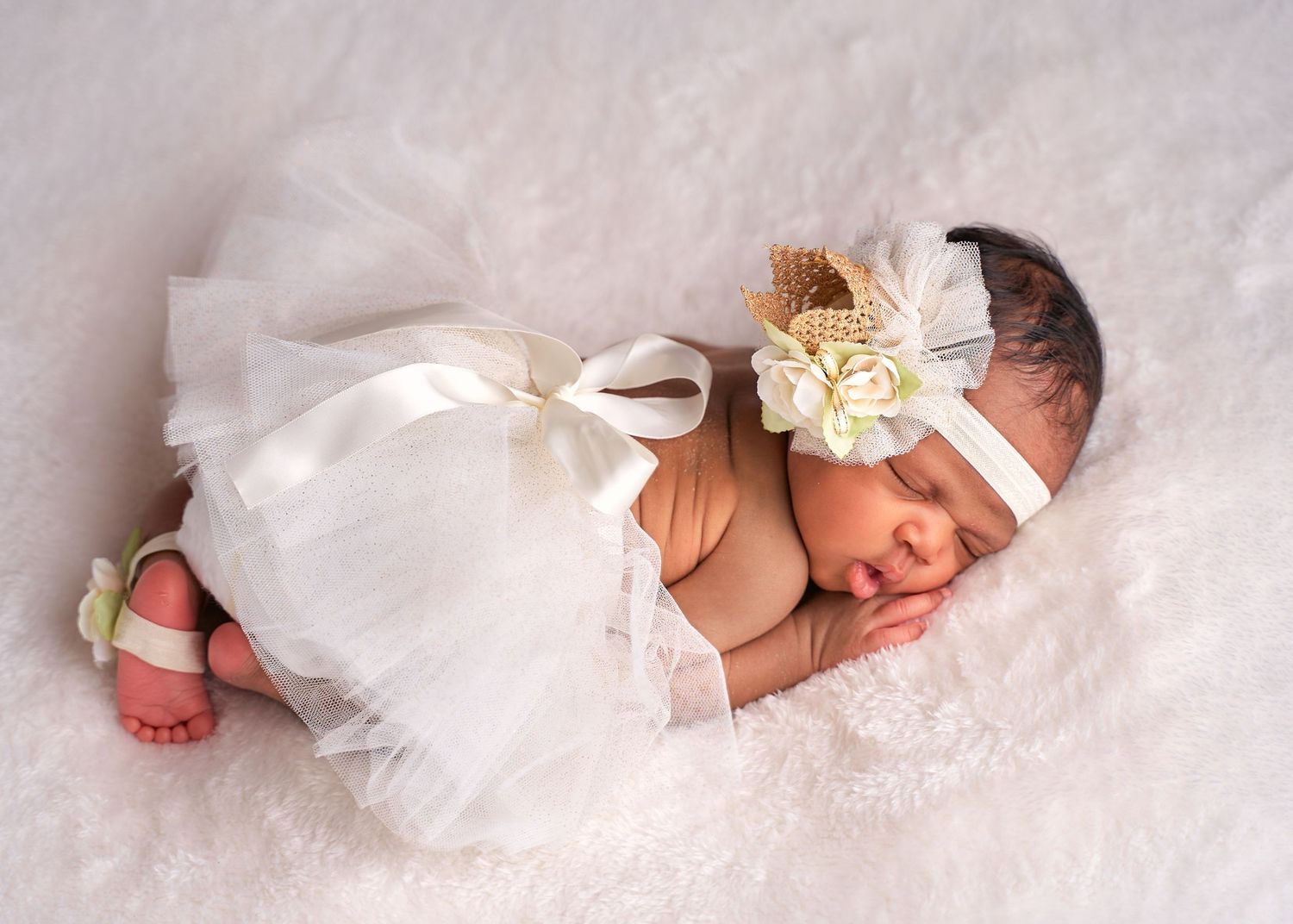 A sleeping newborn baby wearing a white headband lies on soft white fabric in a peaceful pose.