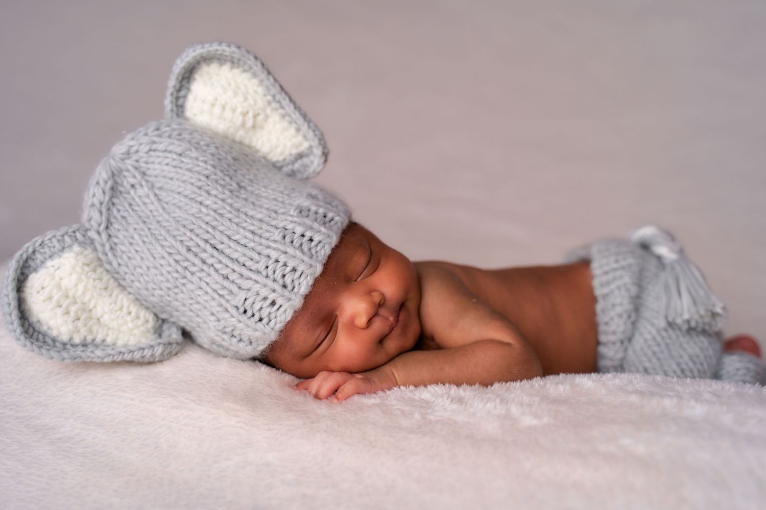 Adorable newborn baby wearing a gray knitted bunny hat lies on soft white bedding.