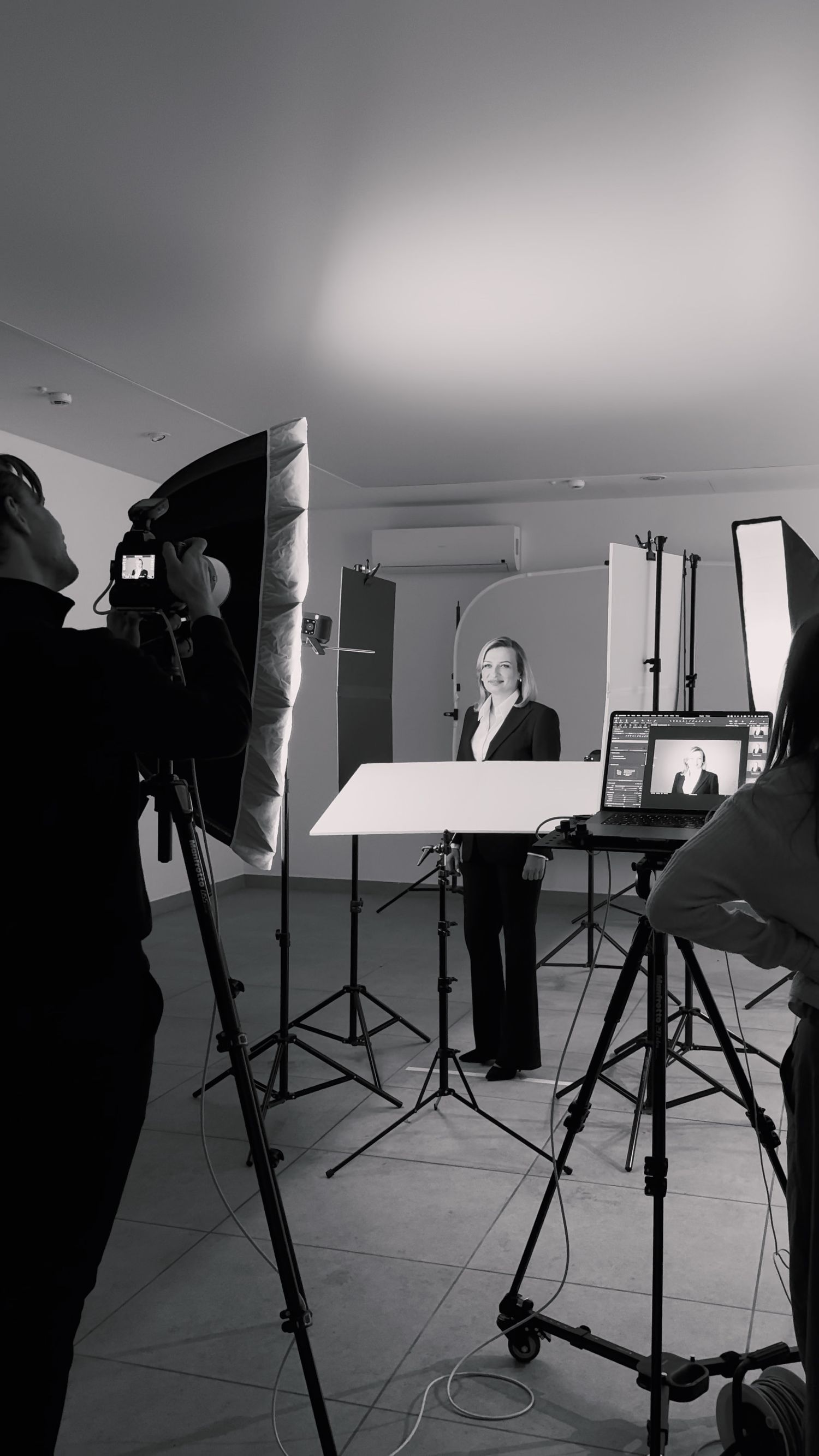 Black and white photo of a professional photo studio shoot with softbox lights, camera operator, and subject posing against backdrop.