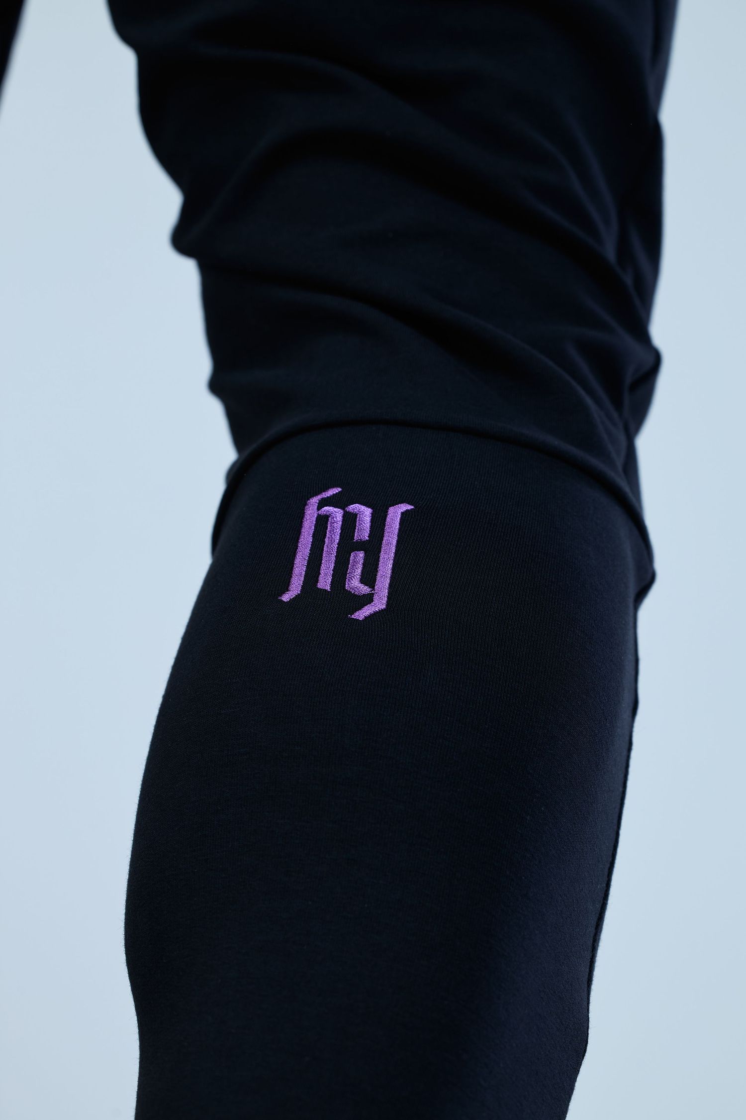 Black leggings with purple embroidered HY logo detail on thigh, worn by a person against a light blue background.