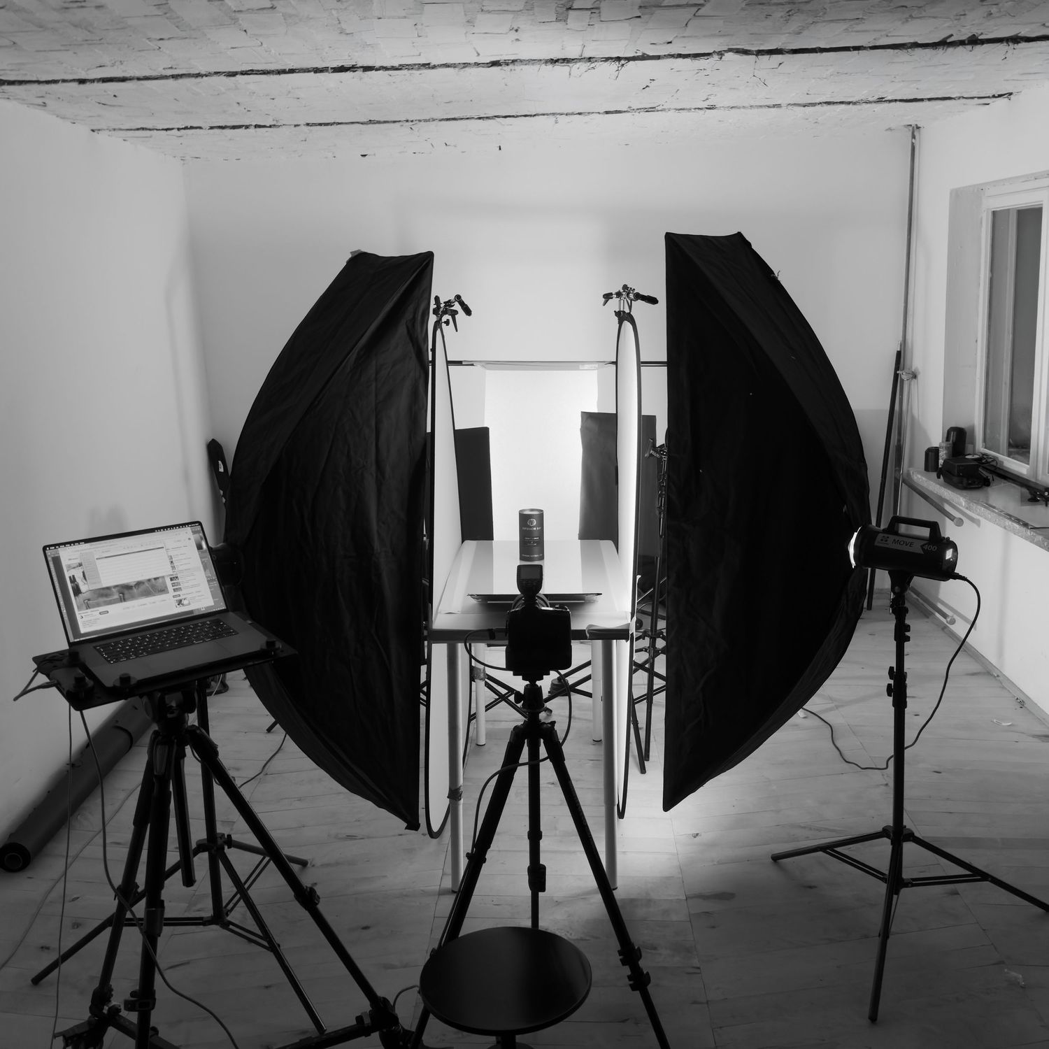 Black and white photo of a professional photography studio setup with softboxes, tripods, and a laptop on a white background.