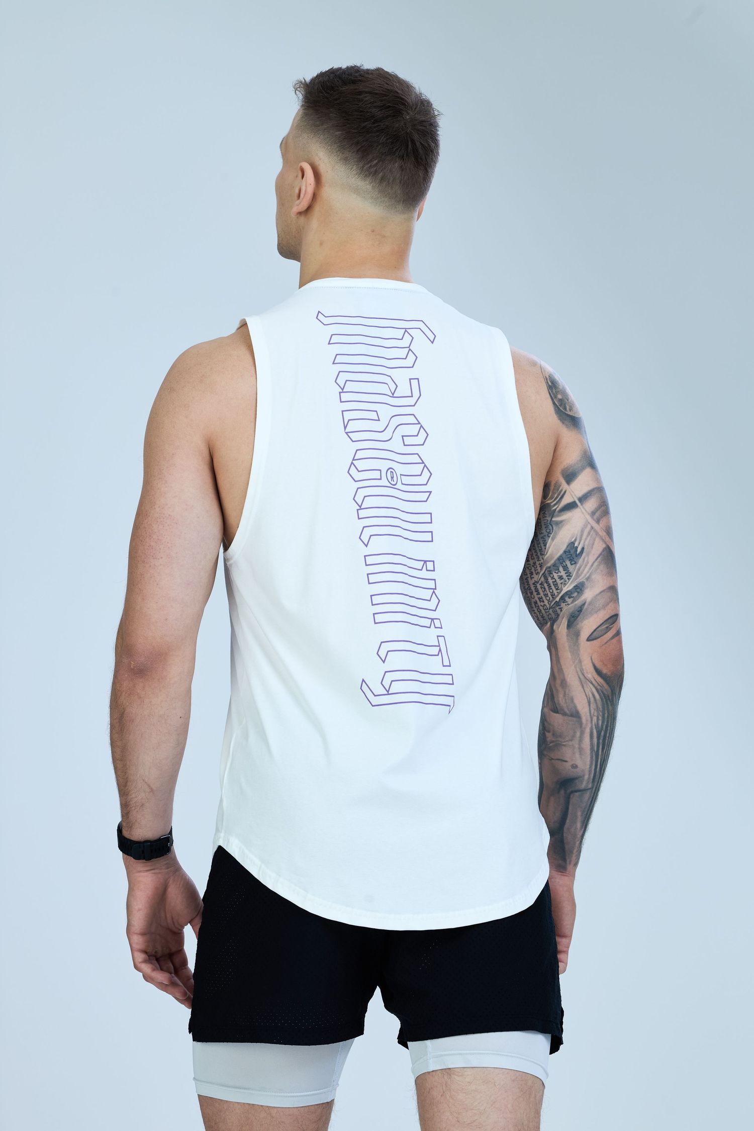 Man wearing white sleeveless tank top with Masculinity print on back, paired with black shorts, showing tattoos.