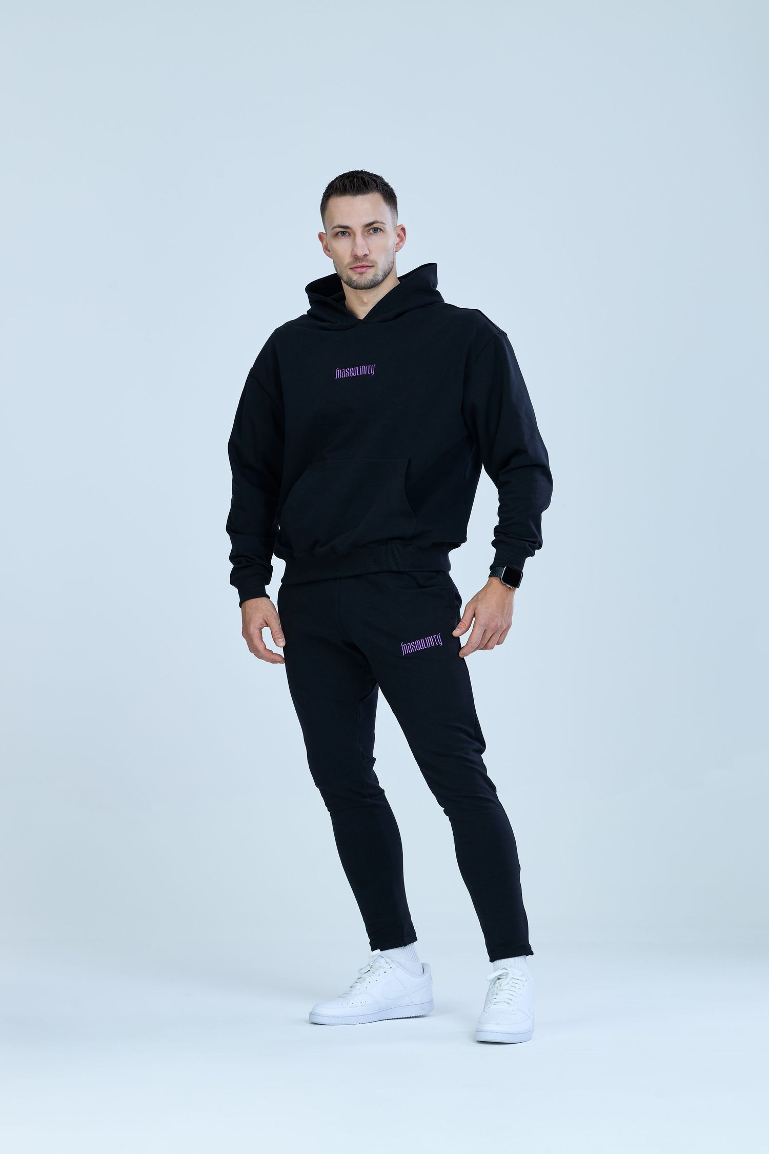 Man wearing black oversized hoodie and slim joggers with white sneakers, full body shot on light background.
