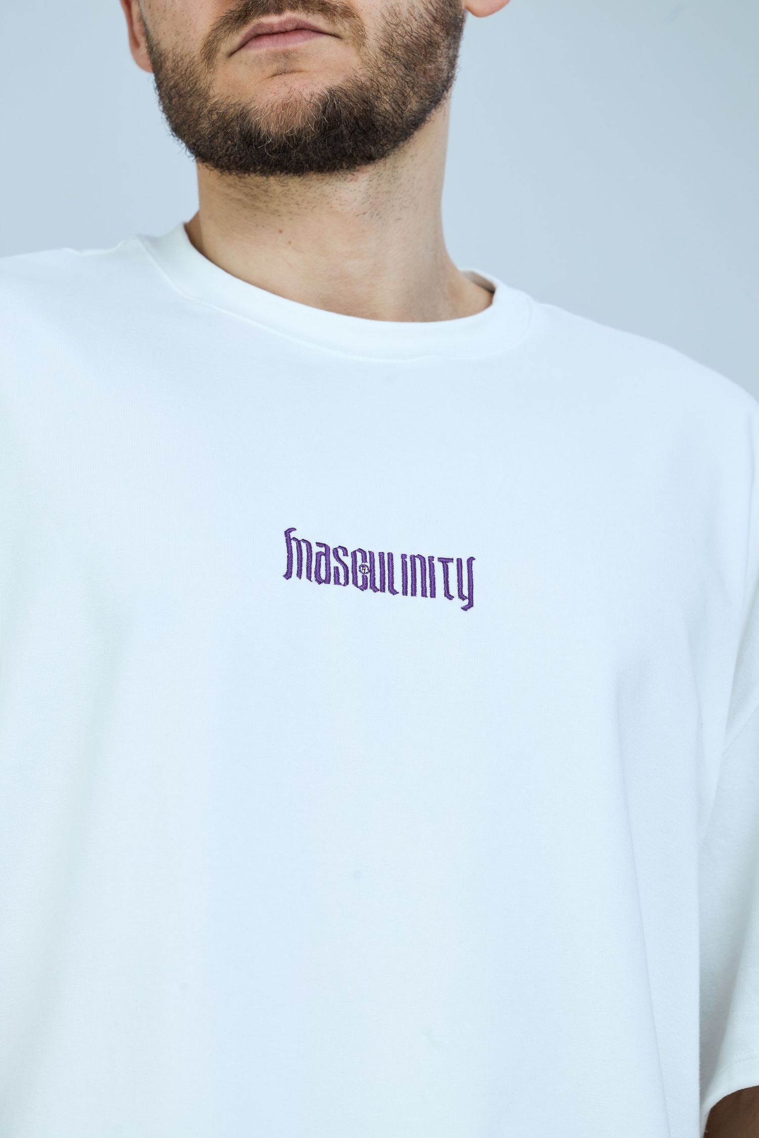 White oversized t-shirt with purple gothic-style 'masculinity' embroidered text on chest, worn by bearded male model.