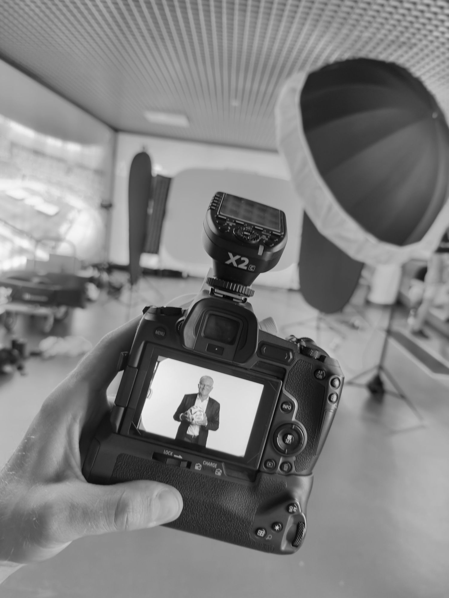 Czarno-białe zdjęcie dłoni trzymającej aparat DSLR, na którego ekranie widać mężczyznę w profesjonalnym studiu fotograficznym.