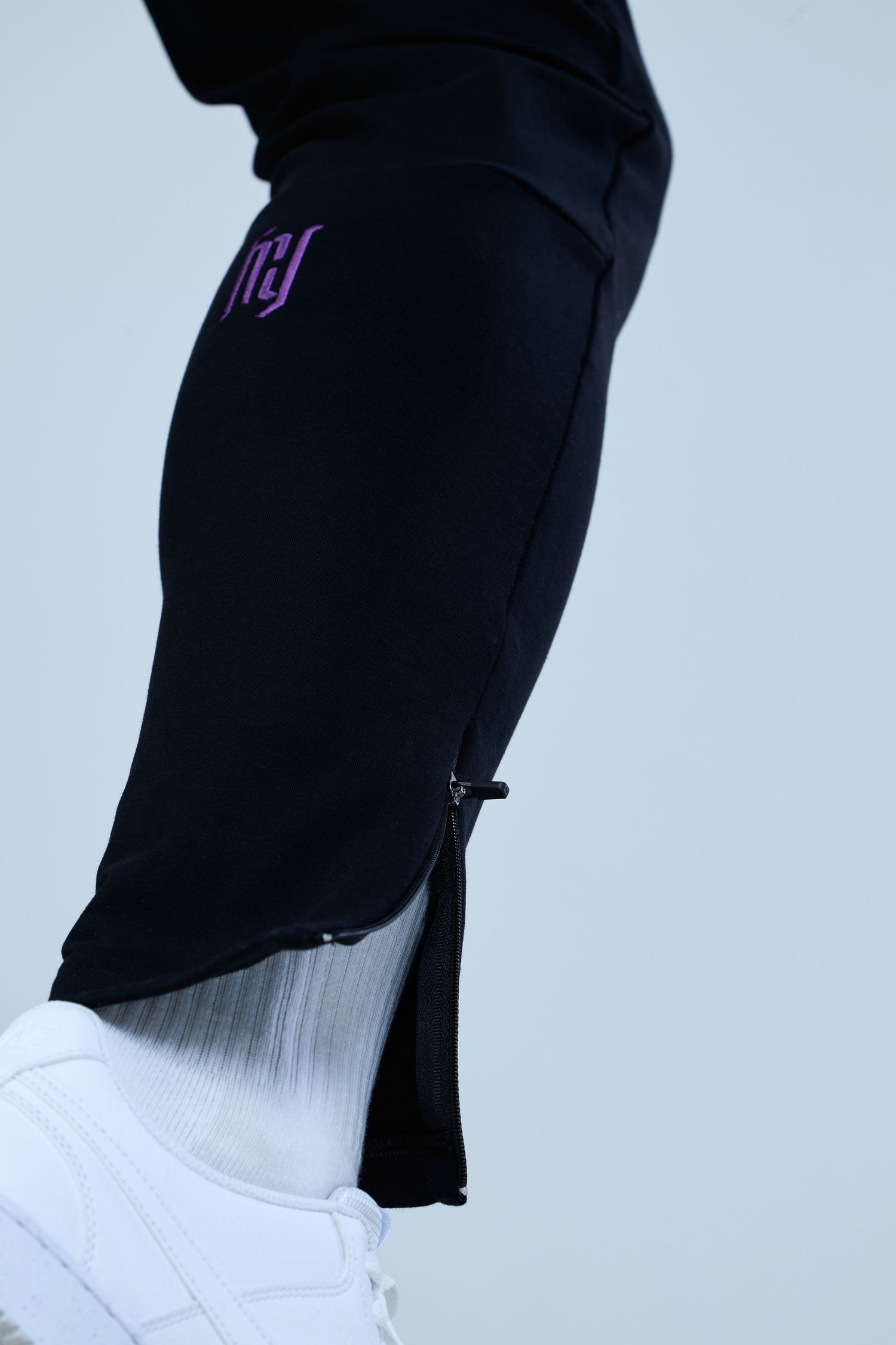 Black jogger pants with purple logo detail and gold zipper hem, paired with white sneakers and socks.