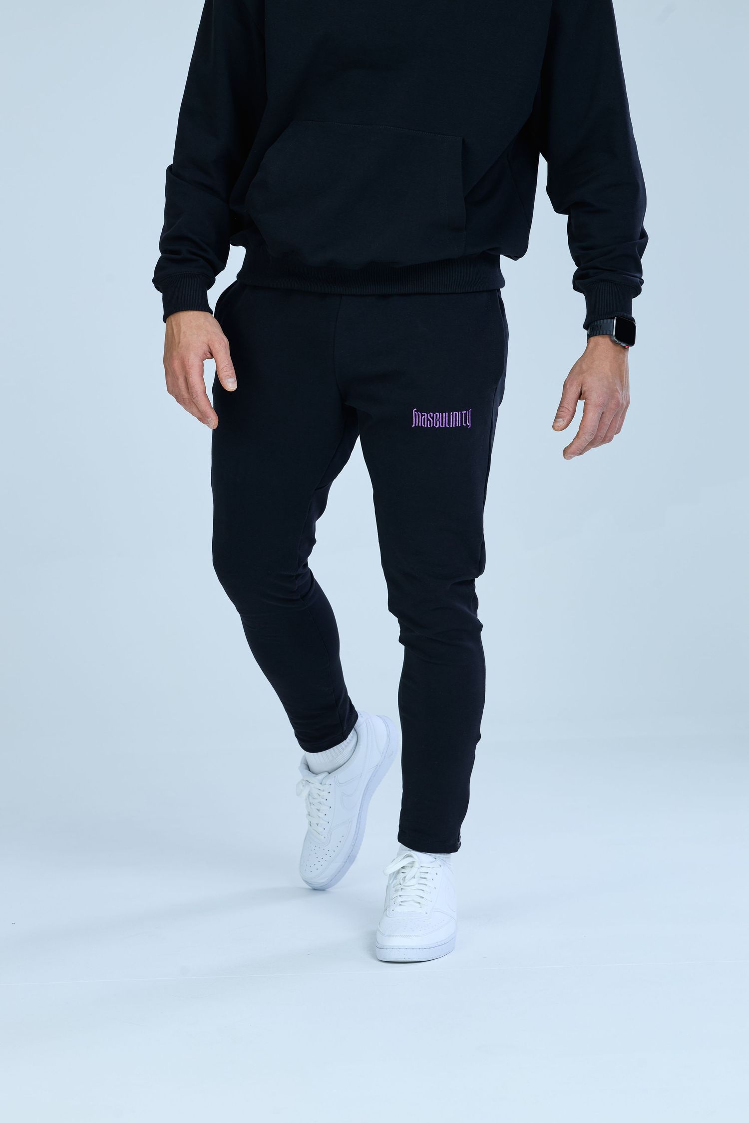 Man wearing black jogger sweatpants with purple logo and matching black hoodie, paired with white sneakers.