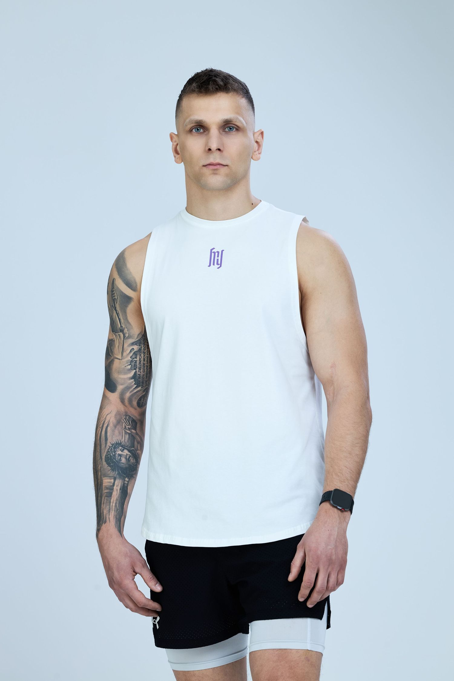 Athletic man wearing a white sleeveless tank top with purple logo and black shorts, showcasing tattoos on arm.