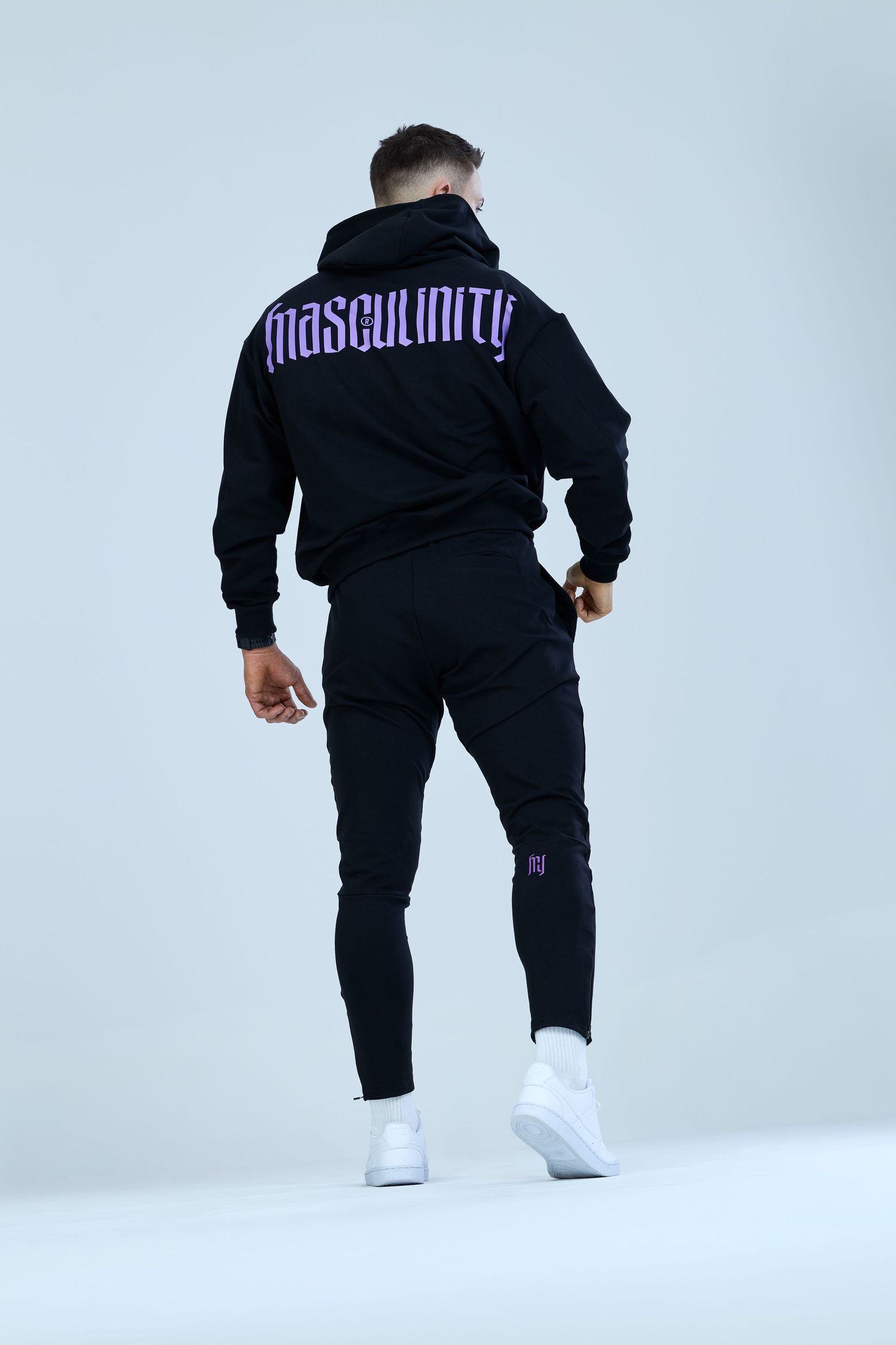 Man wearing black Masculinity hoodie and joggers set with white sneakers, rear view on light background.