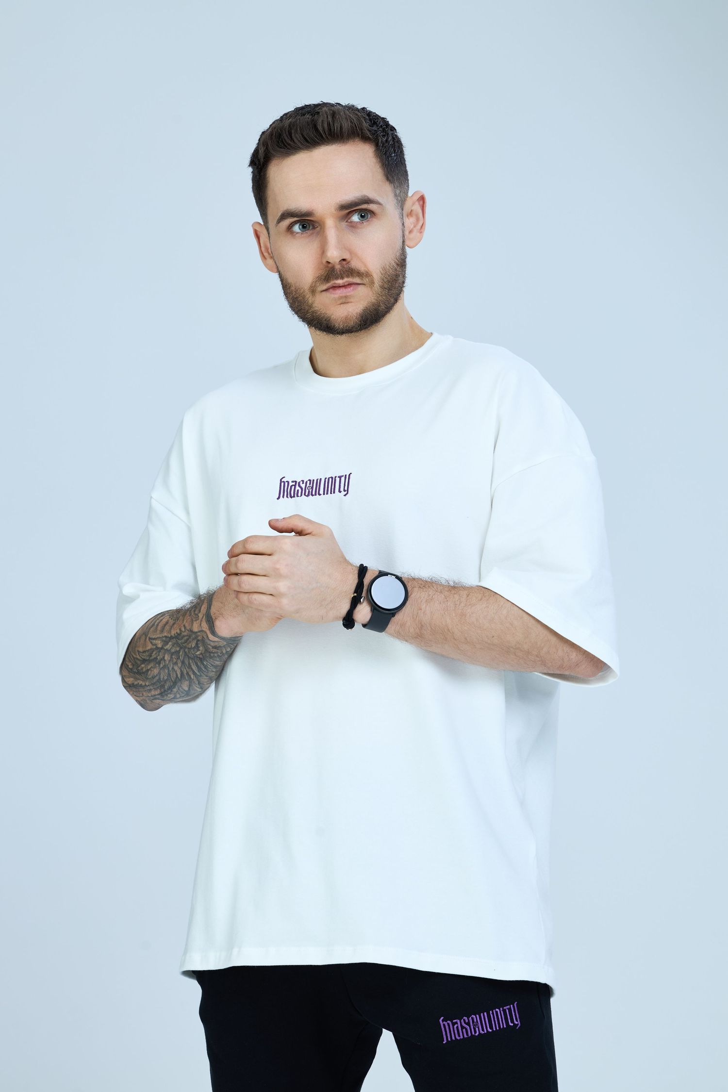 Man wearing oversized white graphic t-shirt with purple logo, black watch, tattoos, and black joggers on light background.