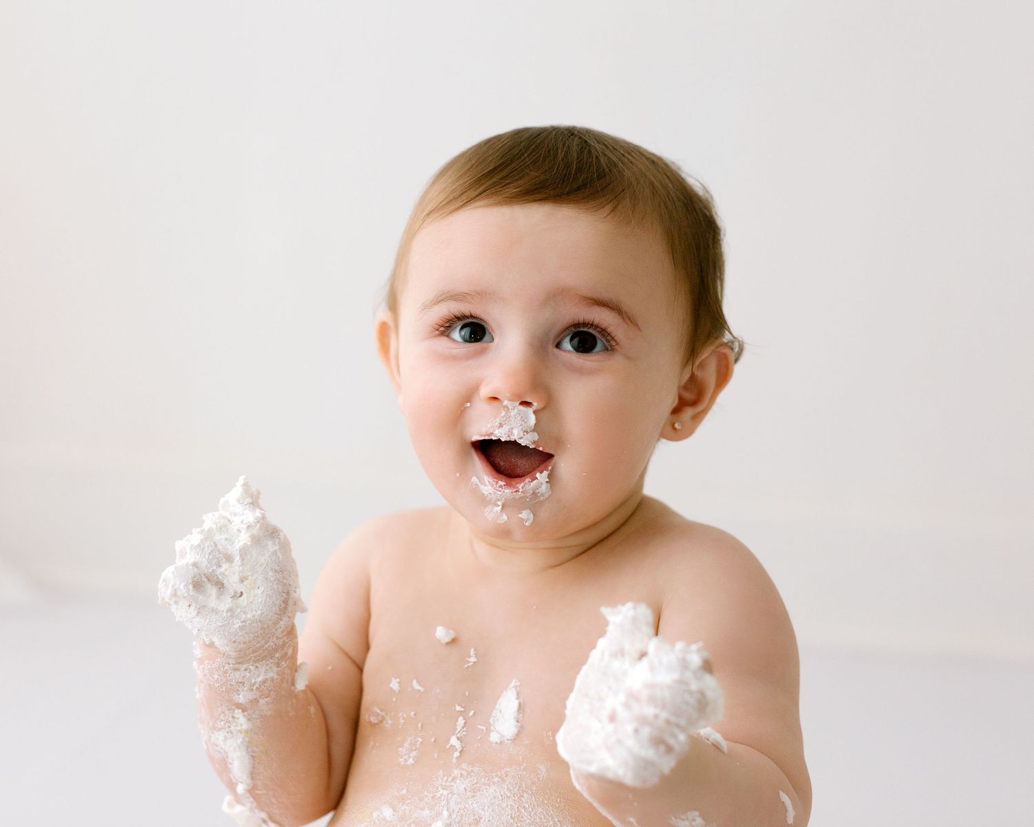 A baby wearing a diaper has cake frosting smeared on their face and hands while sitting against a white background.