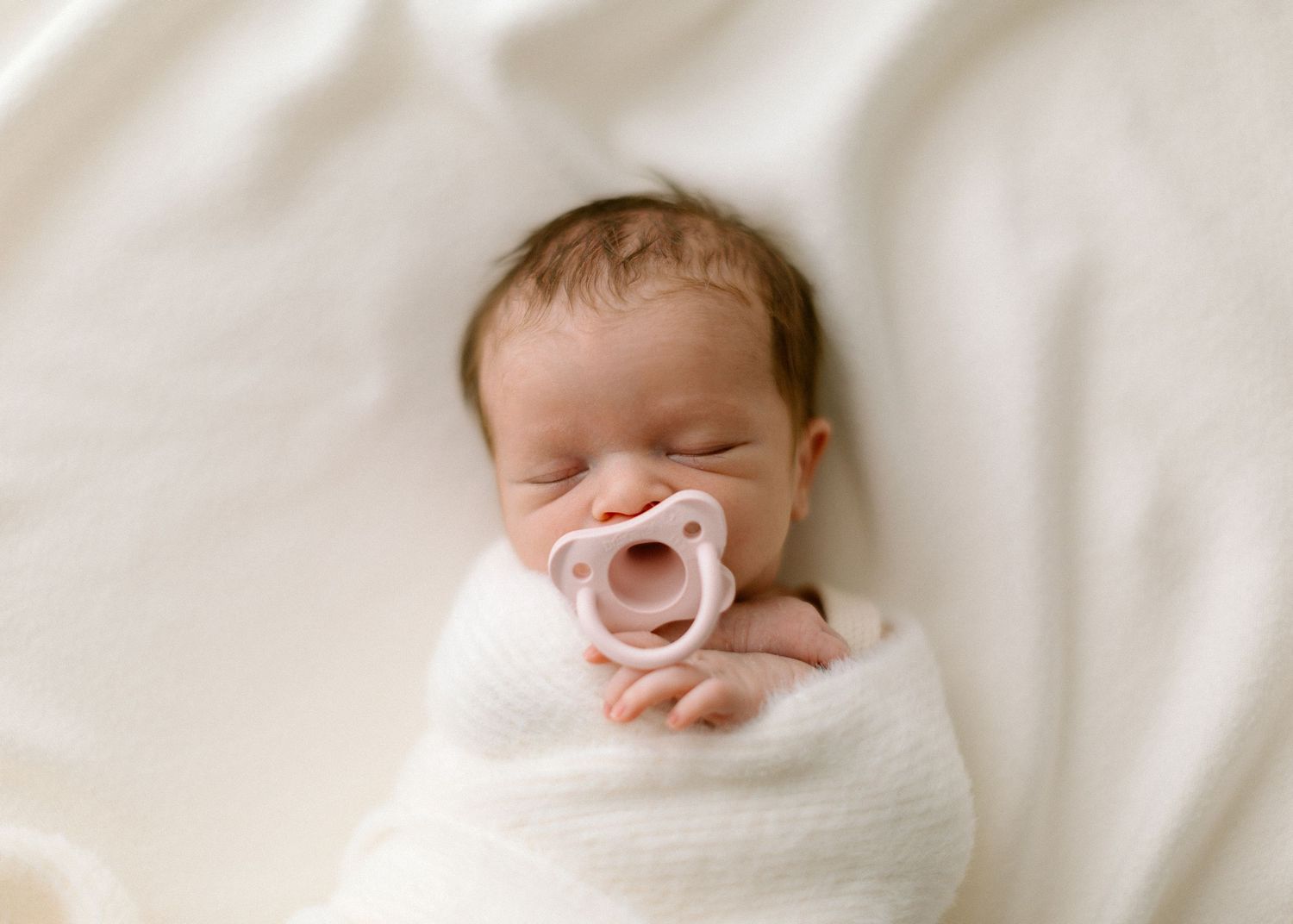 A sleeping newborn baby wrapped in white swaddle blankets rests peacefully on soft bedding.