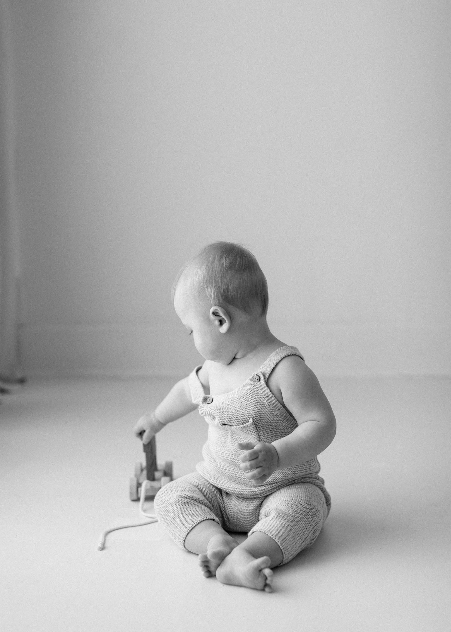 A small child in white overalls sits on the floor playing with a toy in a minimalist black and white setting.