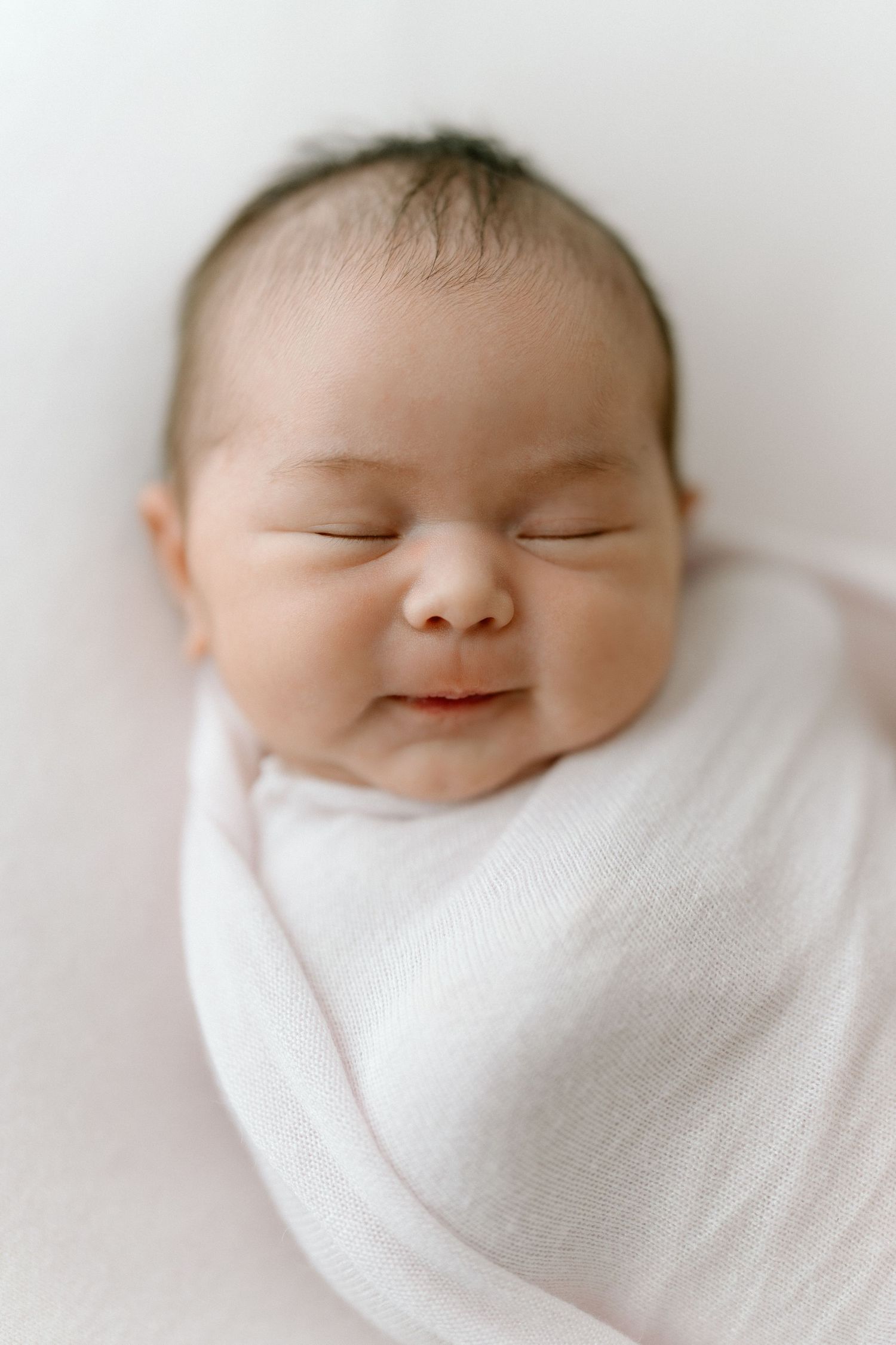 Peaceful sleeping newborn baby wrapped in soft white blanket showing a gentle smile.