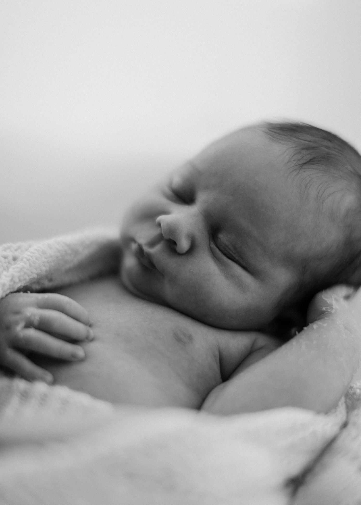 A peaceful black and white photograph shows a sleeping newborn baby resting on soft white bedding.