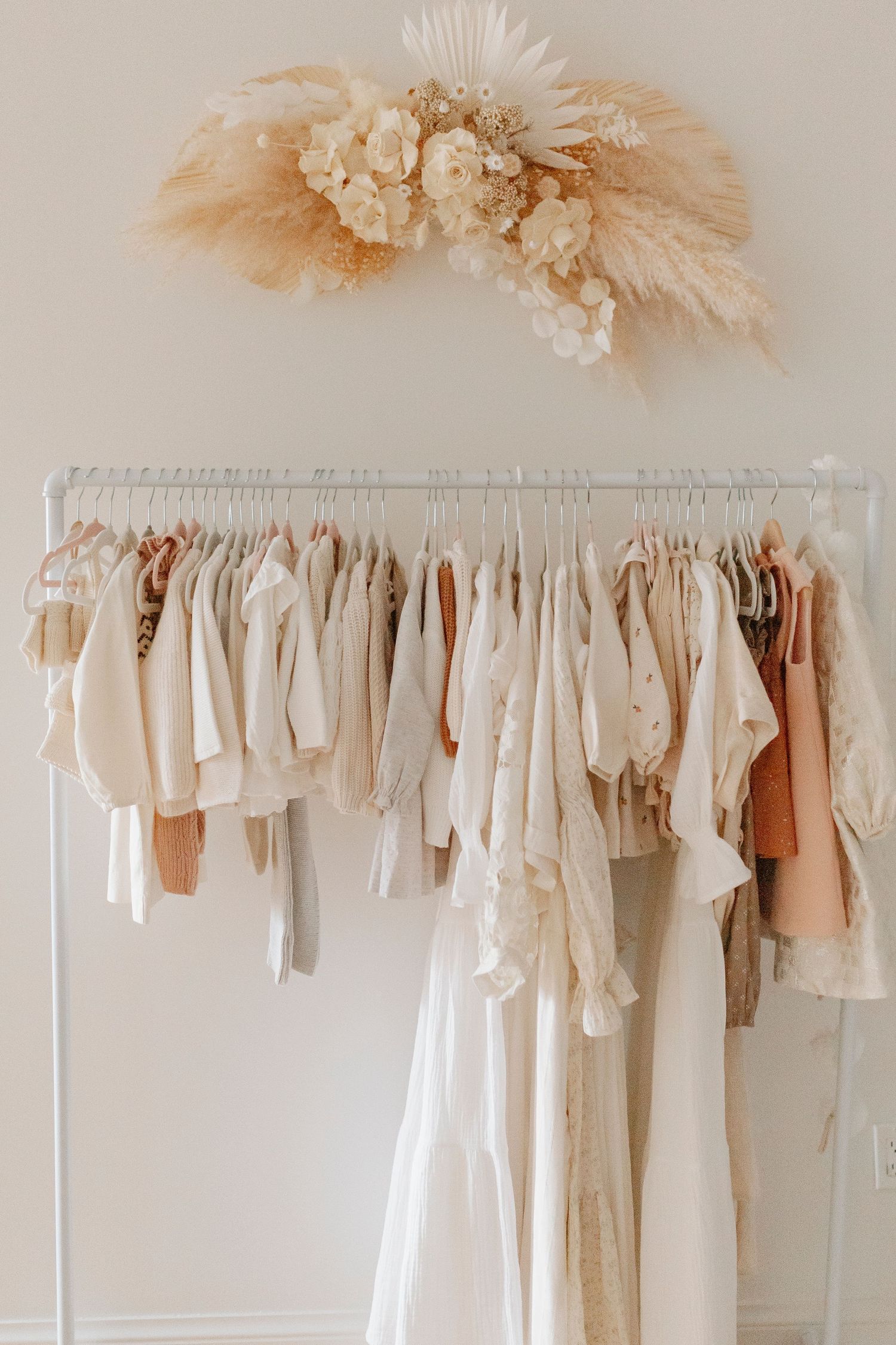 A clothing rack with neutral colored baby clothes and dried floral decoration above.