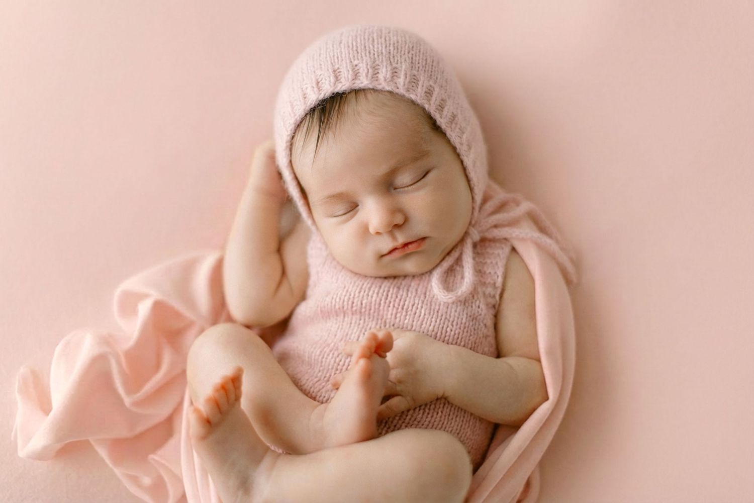 Sleeping newborn baby wearing pink bonnet and wrapped in soft pink fabric on neutral backdrop.