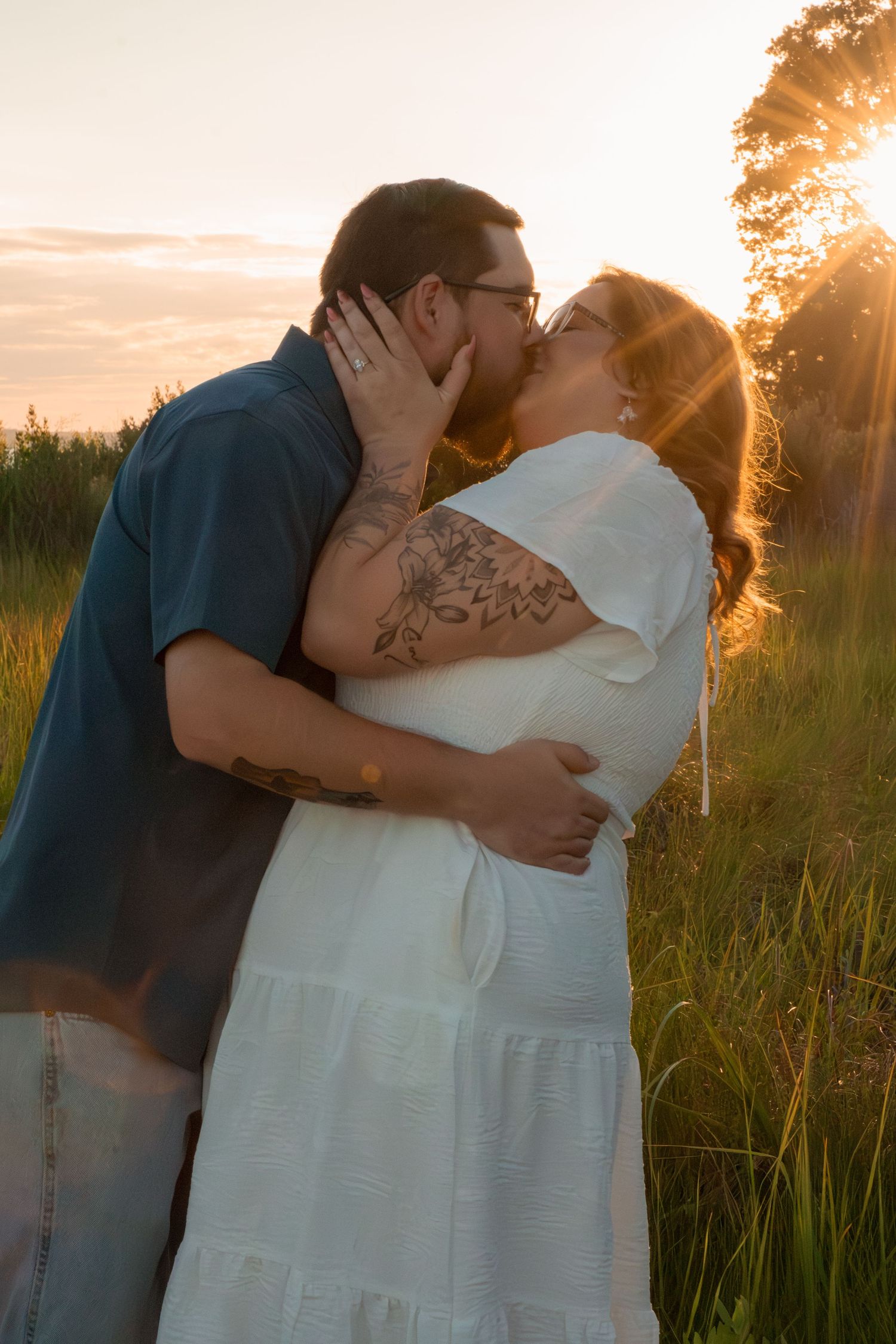Couple sharing a romantic kiss at golden hour sunset in a green field, woman in white dress with floral tattoo.
