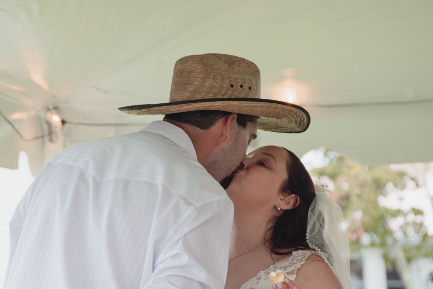 A couple in cowboy hats share a romantic kiss while standing outdoors in bright sunlight.
