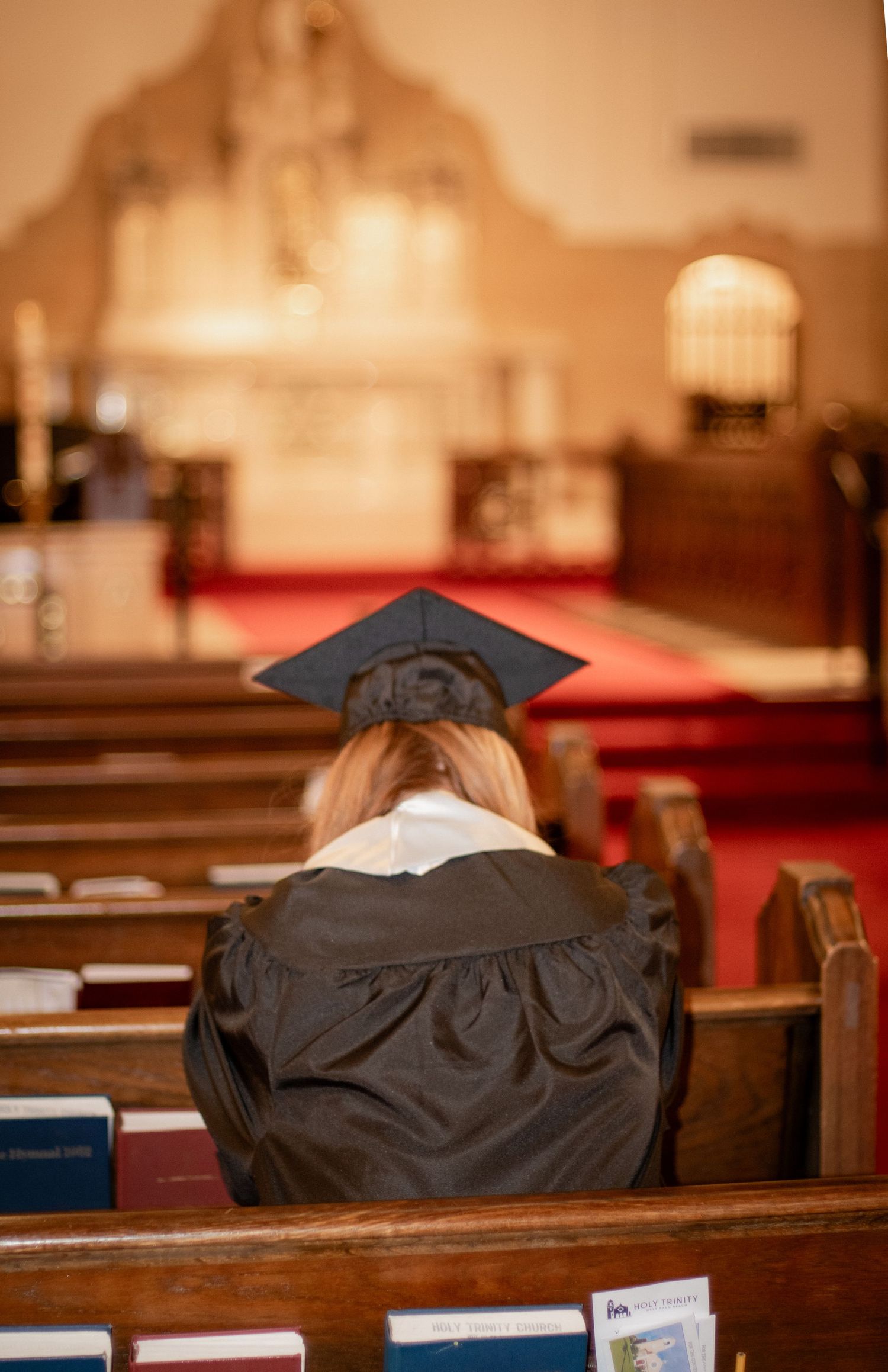 Graduate in cap and gown sitting alone in church pew, bowing head in prayer before altar.