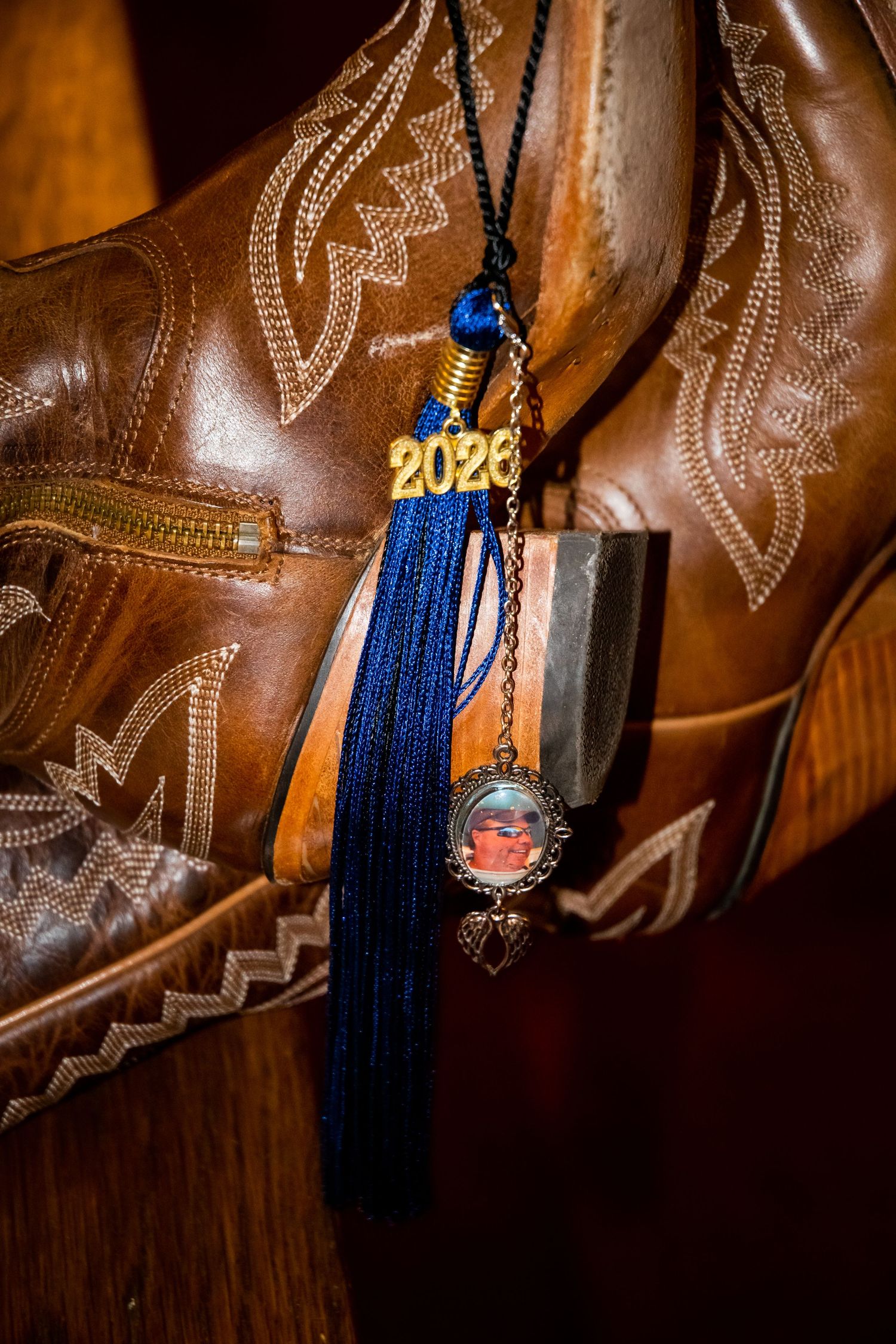 Class of 2026 graduation tassel with blue fringe and locket charm displayed on brown leather cowboy boots.