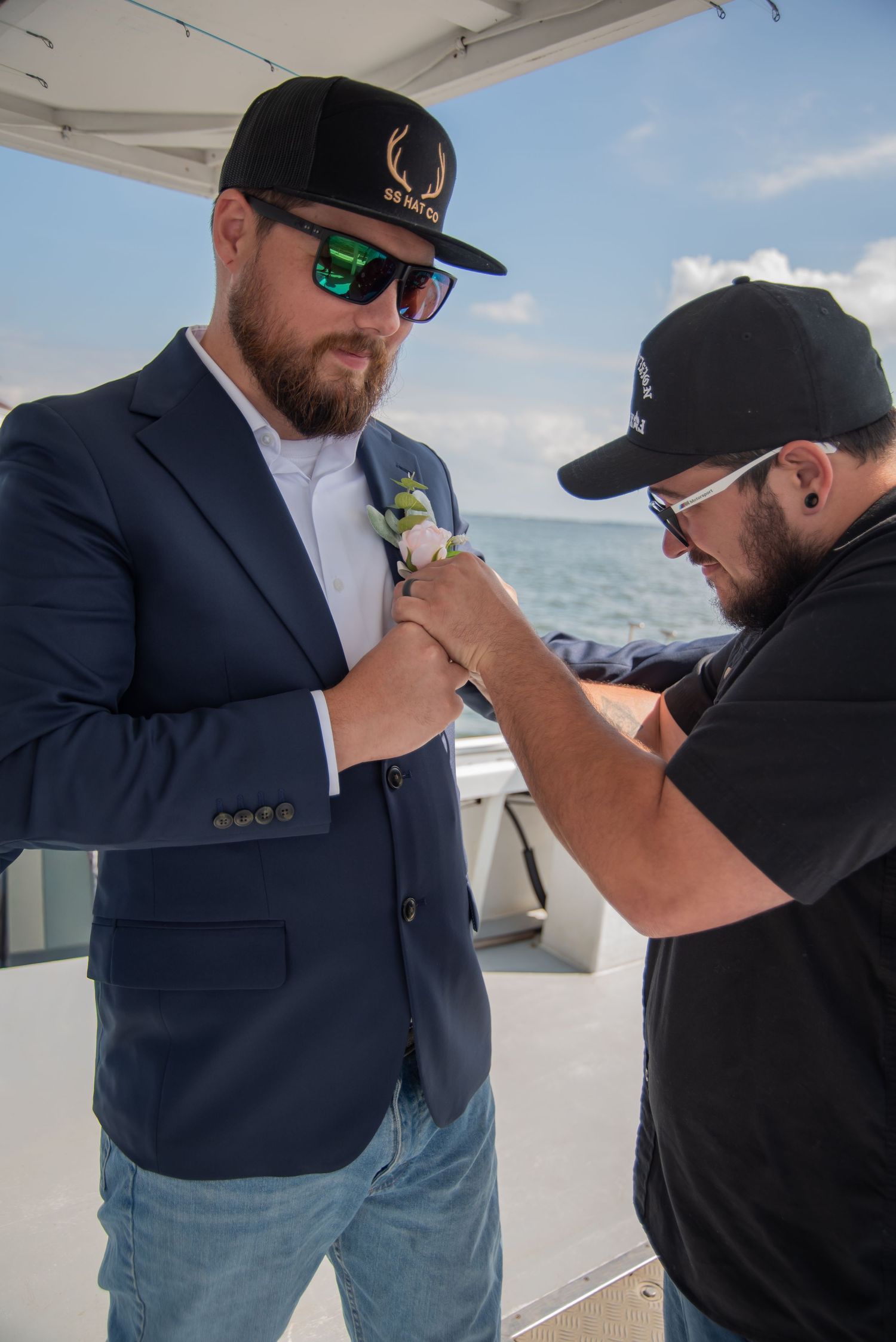 Groom in navy suit gets boutonniere pinned on boat deck with ocean backdrop.