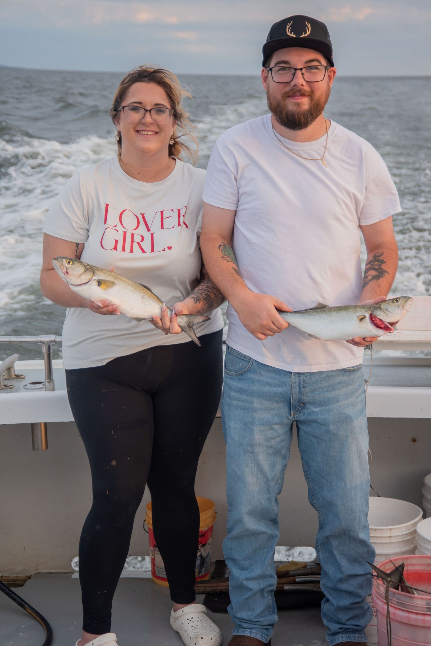A couple proudly holds freshly caught fish on a boat deck, woman wearing a Lover Girl shirt, man in jeans and cap.