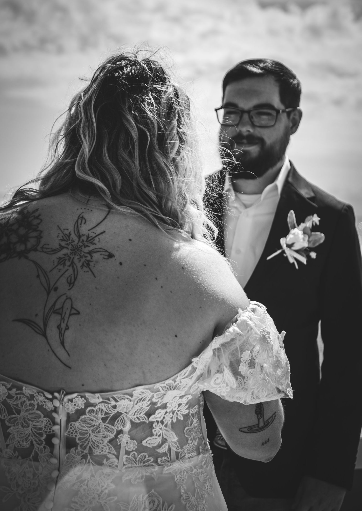 Black and white wedding photo of bride in lace off-shoulder gown with back tattoo facing groom in suit with boutonniere.