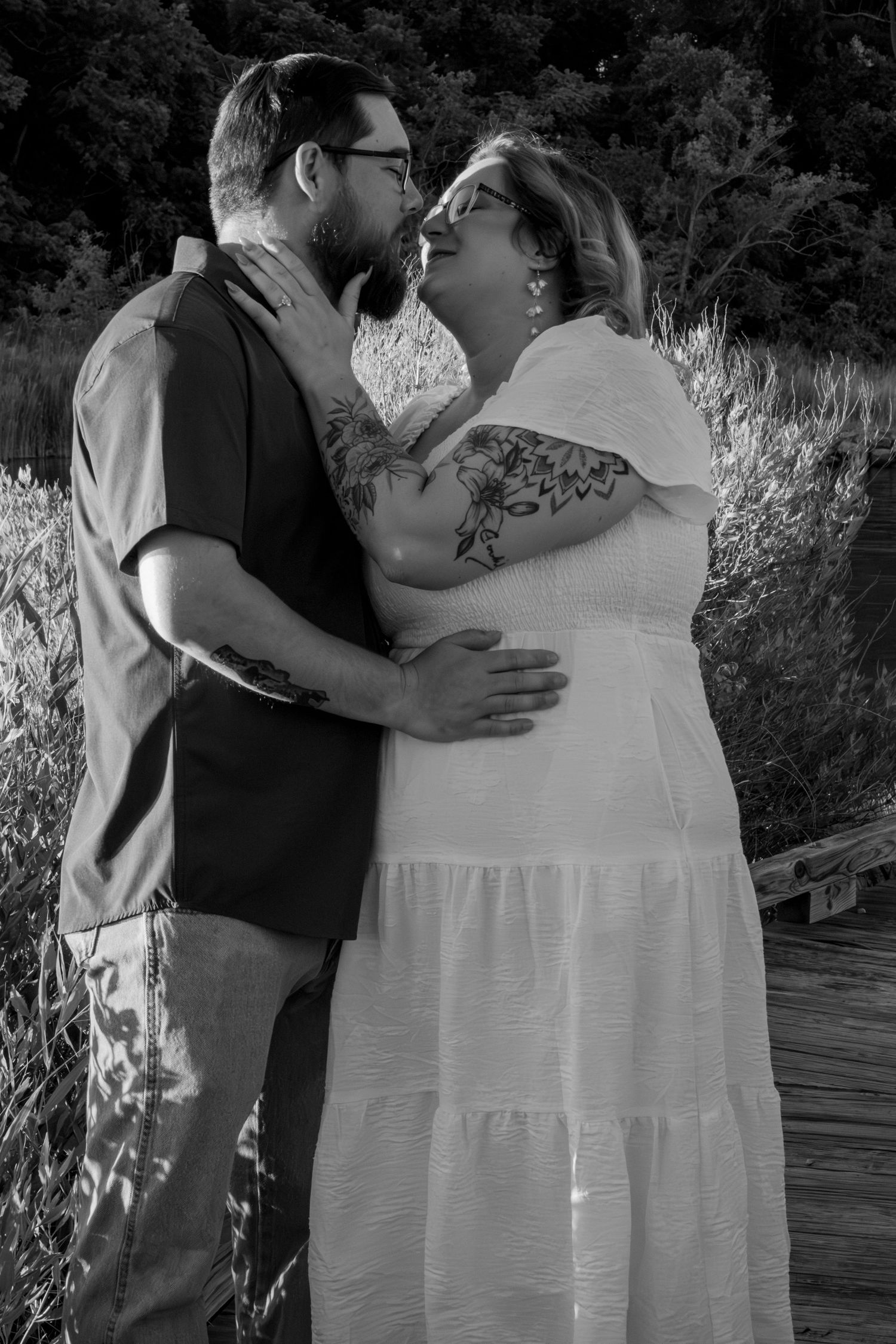 Black and white photo of a couple embracing and kissing outdoors, woman in white dress with tattoos visible.