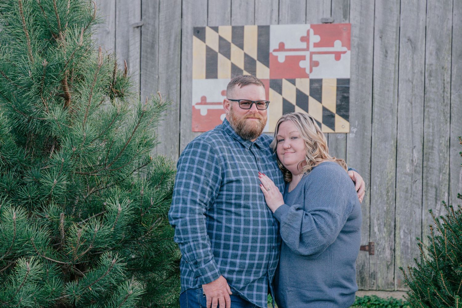 Couple posing by wooden fence and evergreen trees during winter photo session.