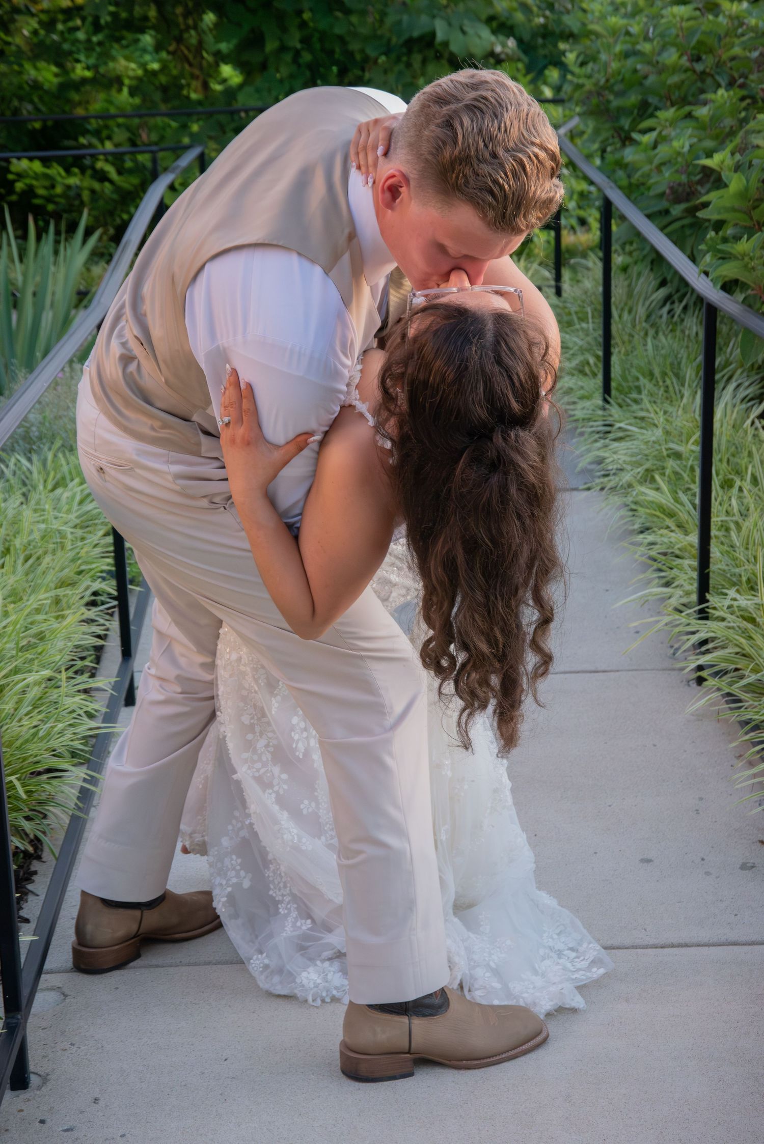 A bride and groom share a romantic dip kiss on a garden bridge, she in lace white gown, he in tan suit with cowboy boots.