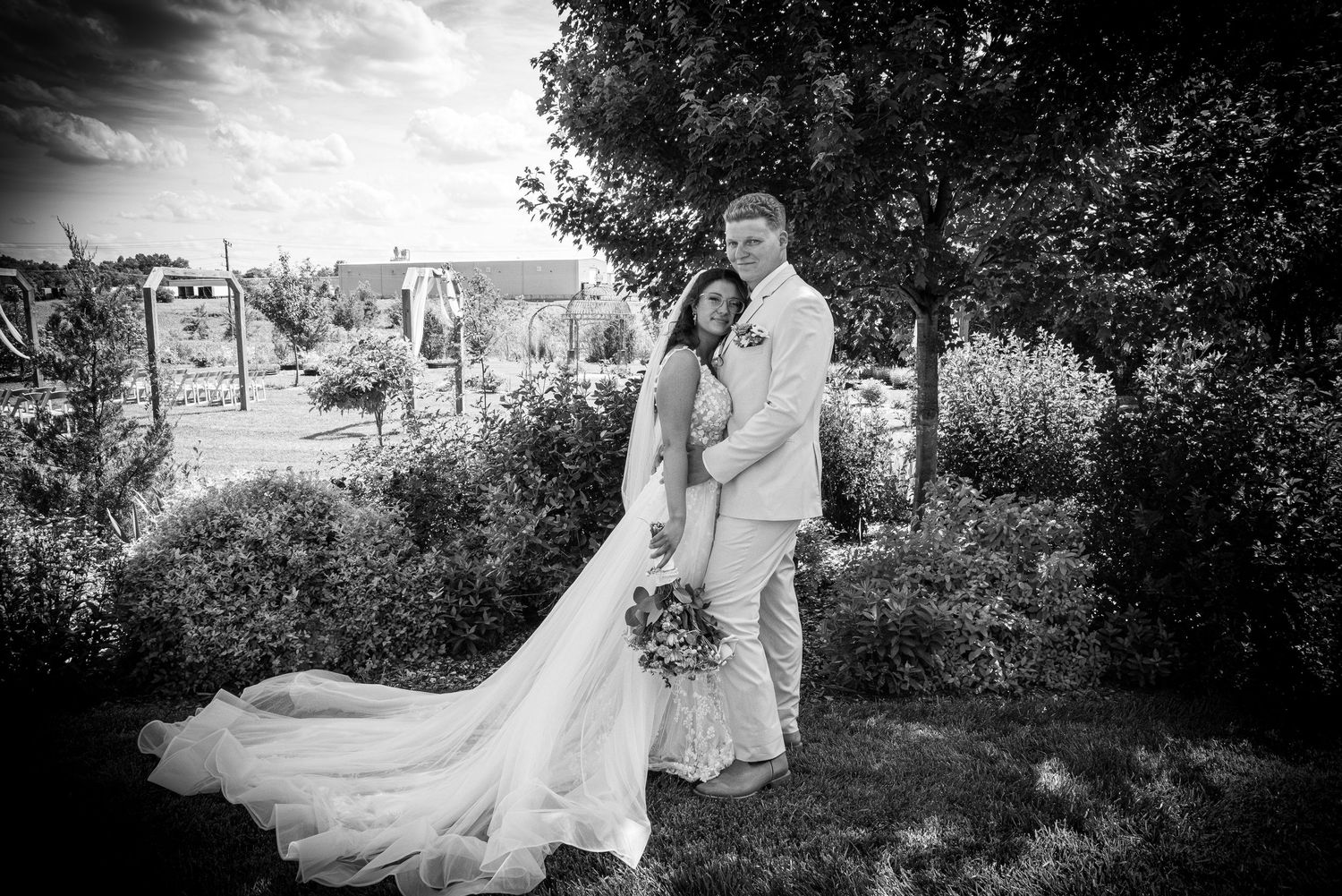 Black and white wedding photo of bride and groom embracing in a lush garden, bride wearing a flowing gown with long train.