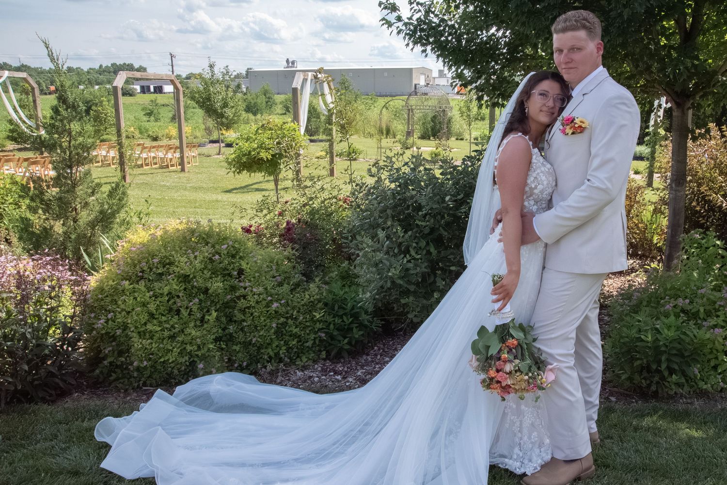 Bride and groom pose in lush garden, bride in long white gown with train, groom in cream suit, holding floral bouquet.