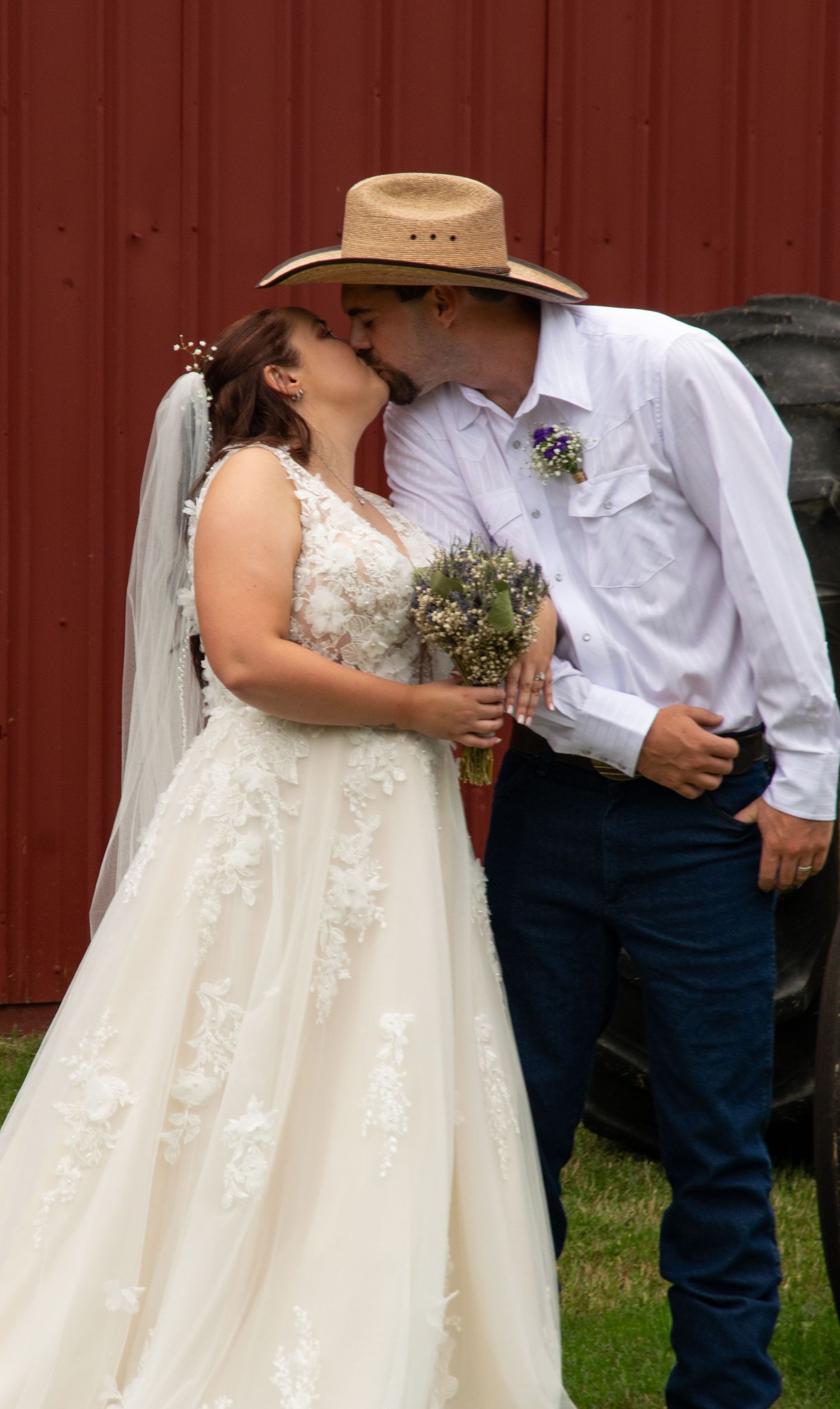 A romantic wedding couple stands against a red barn wall while holding rustic wildflower bouquets.