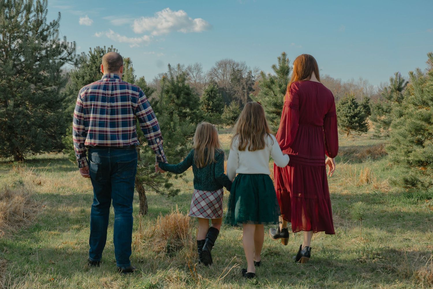 Family walking together through an outdoor field with evergreen trees and blue sky.