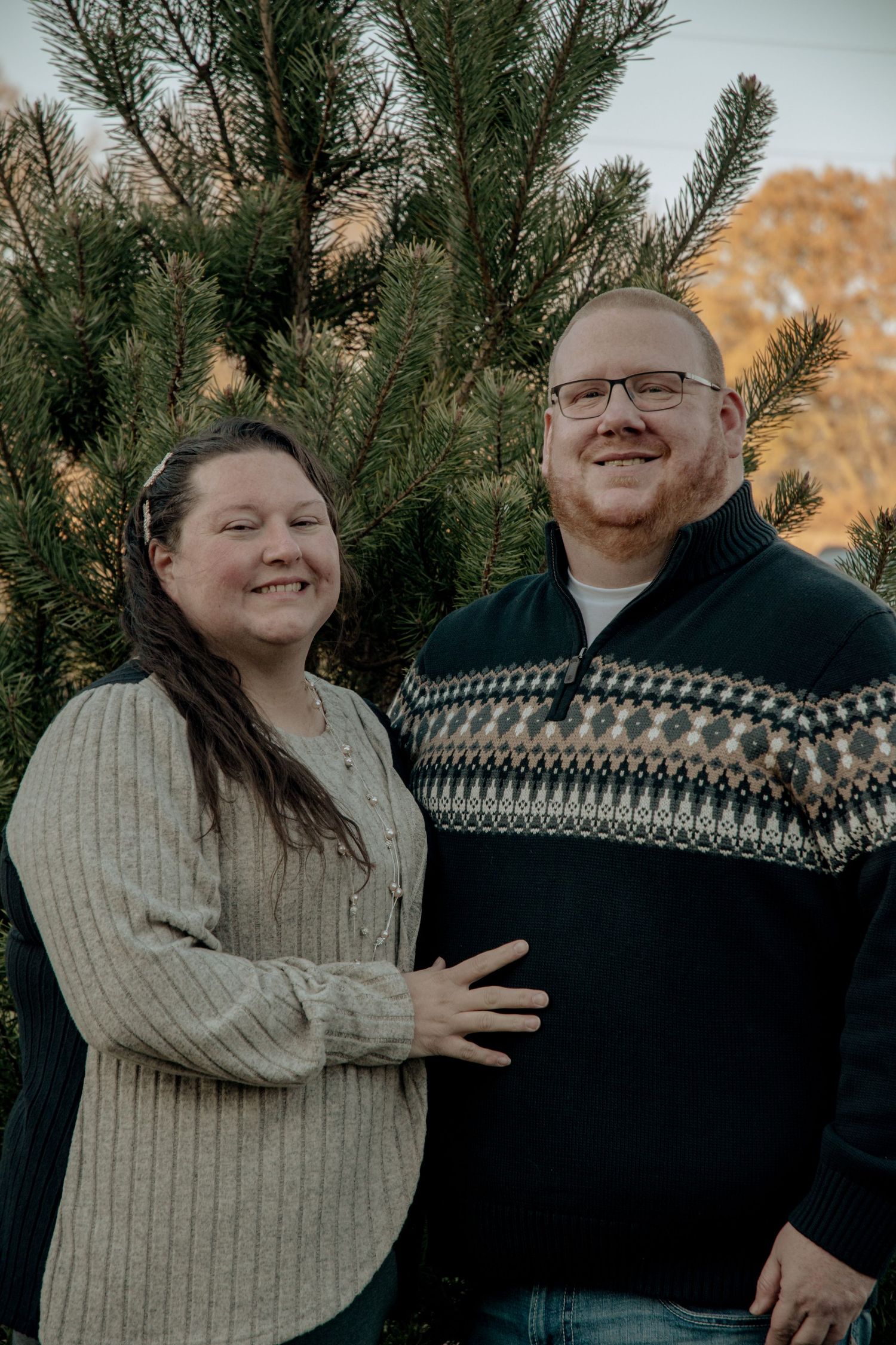 Couple in winter sweaters embracing and laughing near evergreen trees.