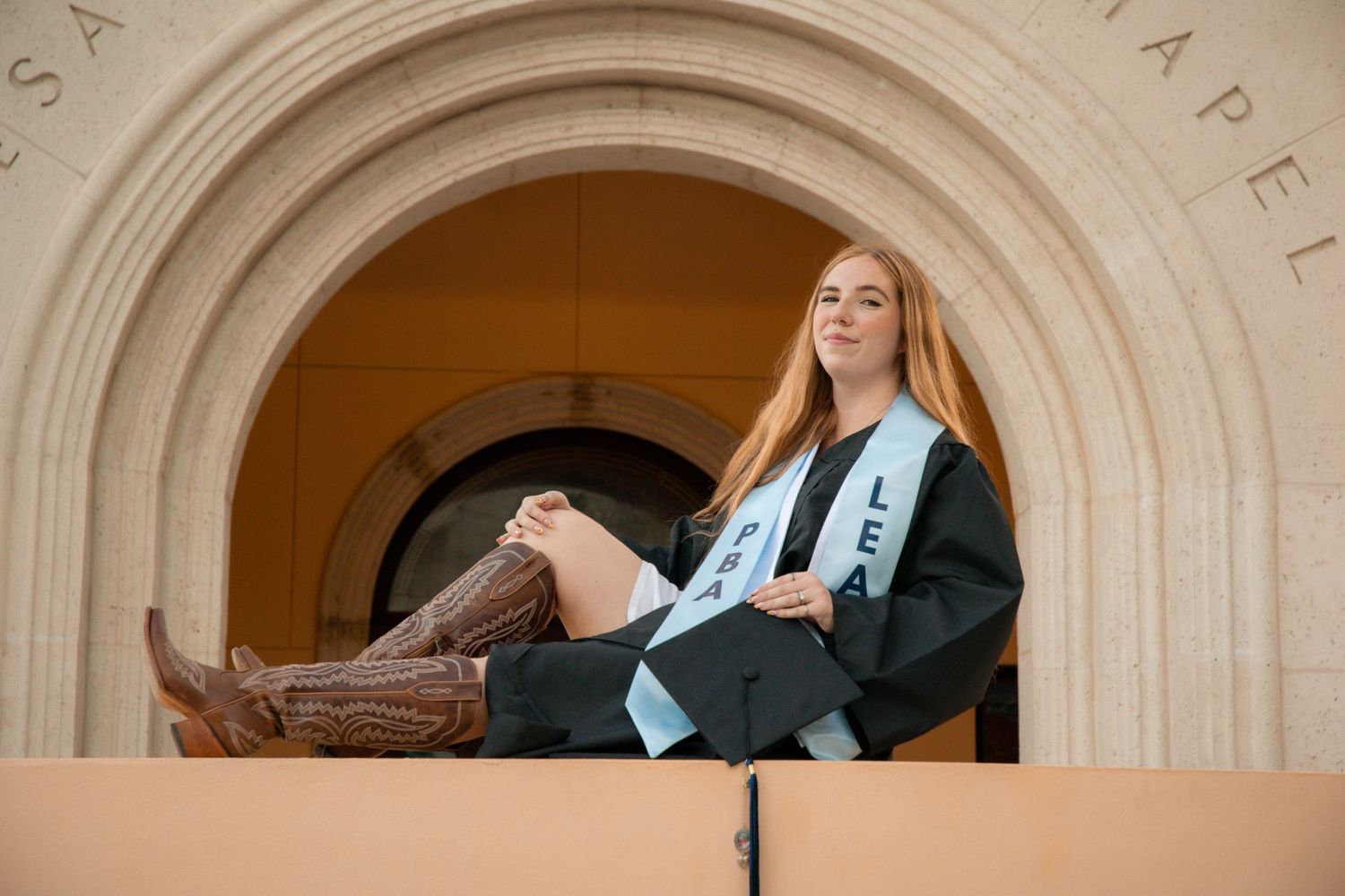 Young woman in graduation cap and gown with sash sits in stone archway wearing cowboy boots at chapel.